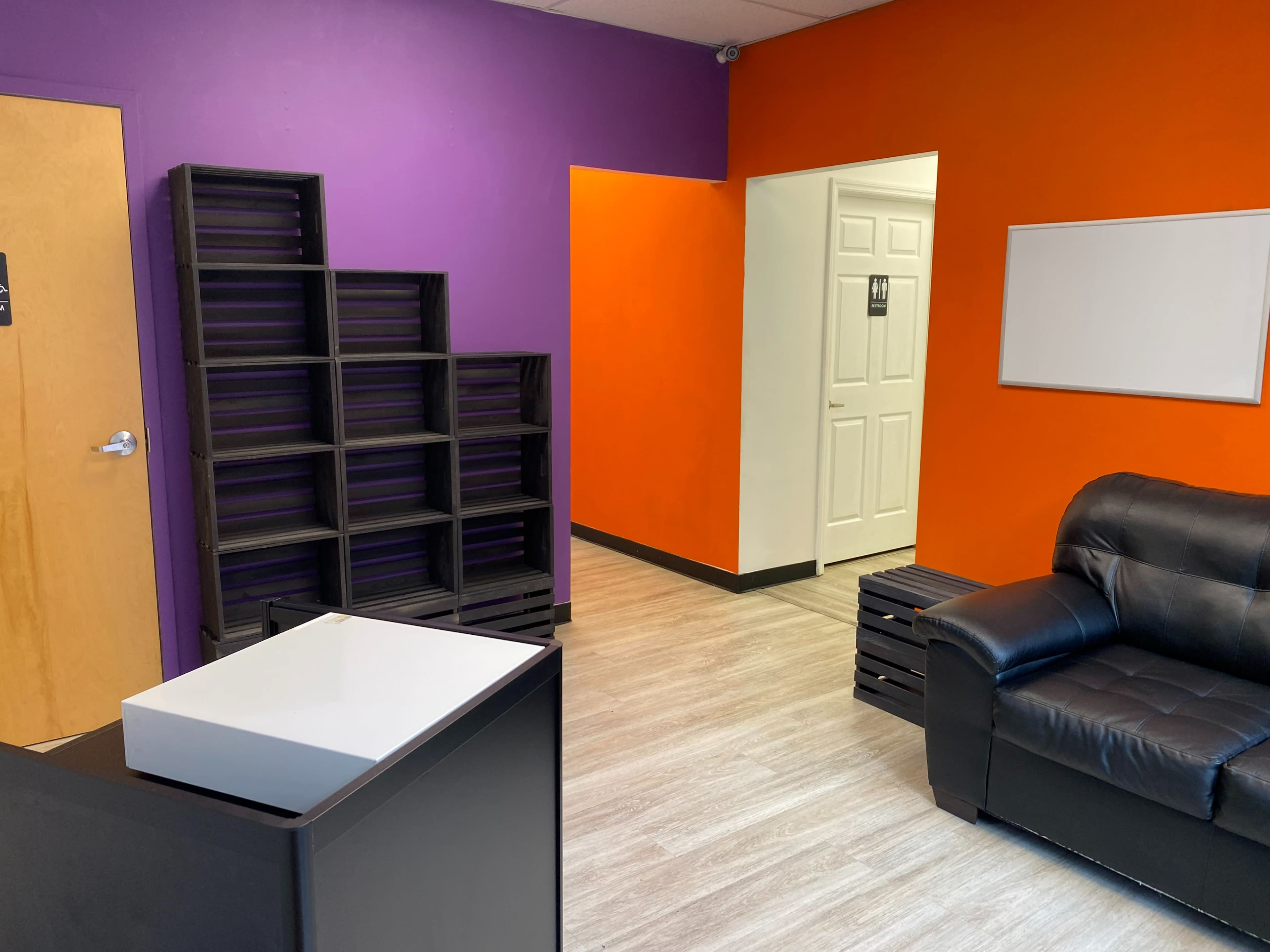 A room with a black sofa, a white table, and a partition featuring purple and orange walls, along with a shelf unit and an entrance to a restroom.