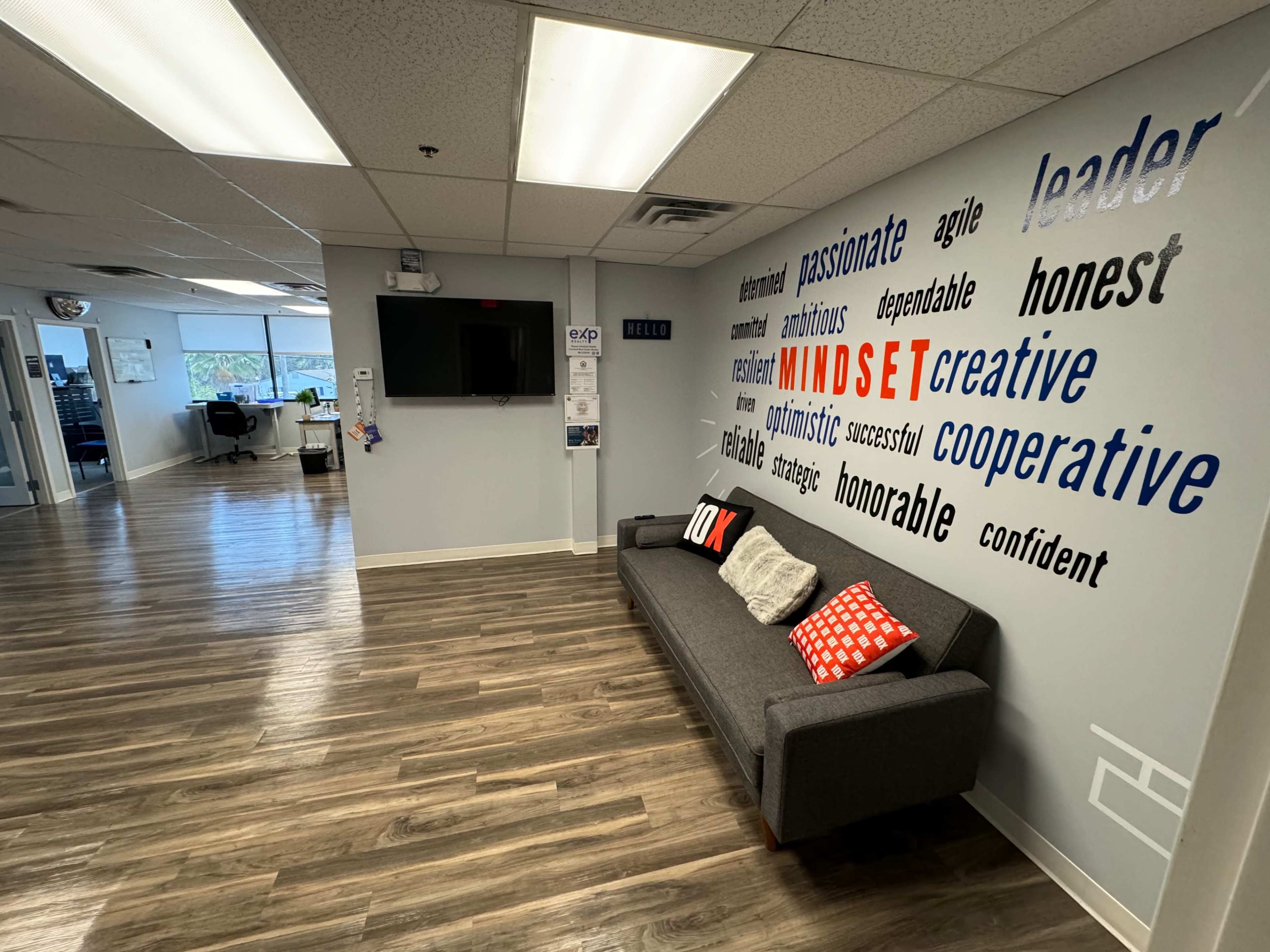 A well-lit office hallway featuring a gray sofa with red and white pillows, and a wall decorated with various positive words related to mindset and leadership.