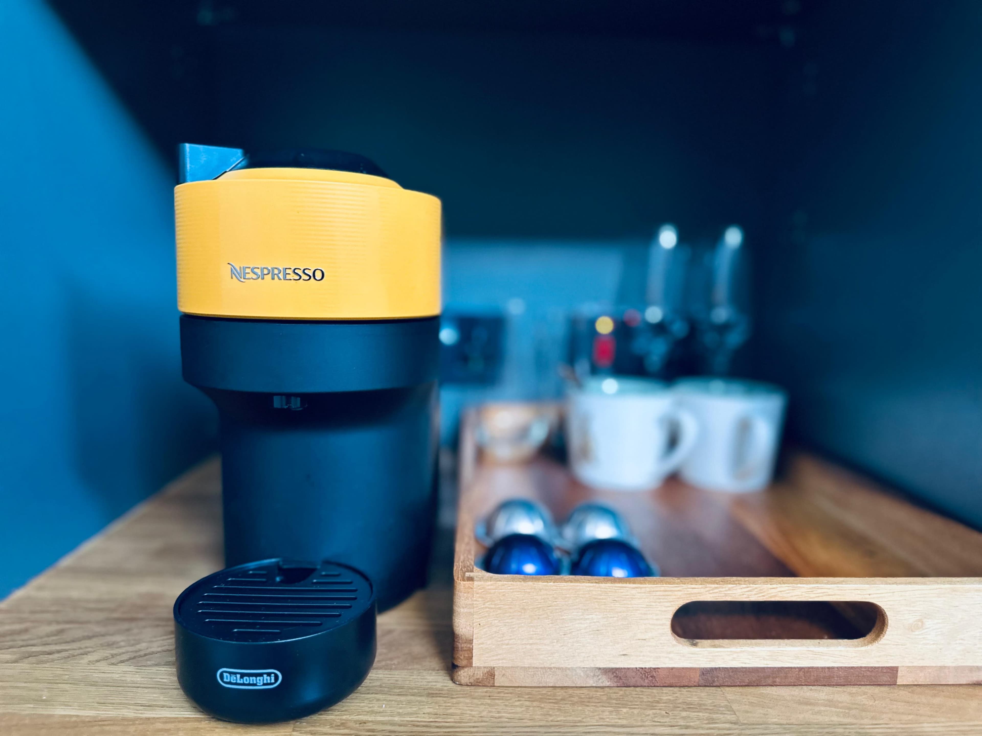 A yellow and black Nespresso coffee maker sits on a wooden tray alongside coffee pods and white mugs in a kitchen cabinet.
