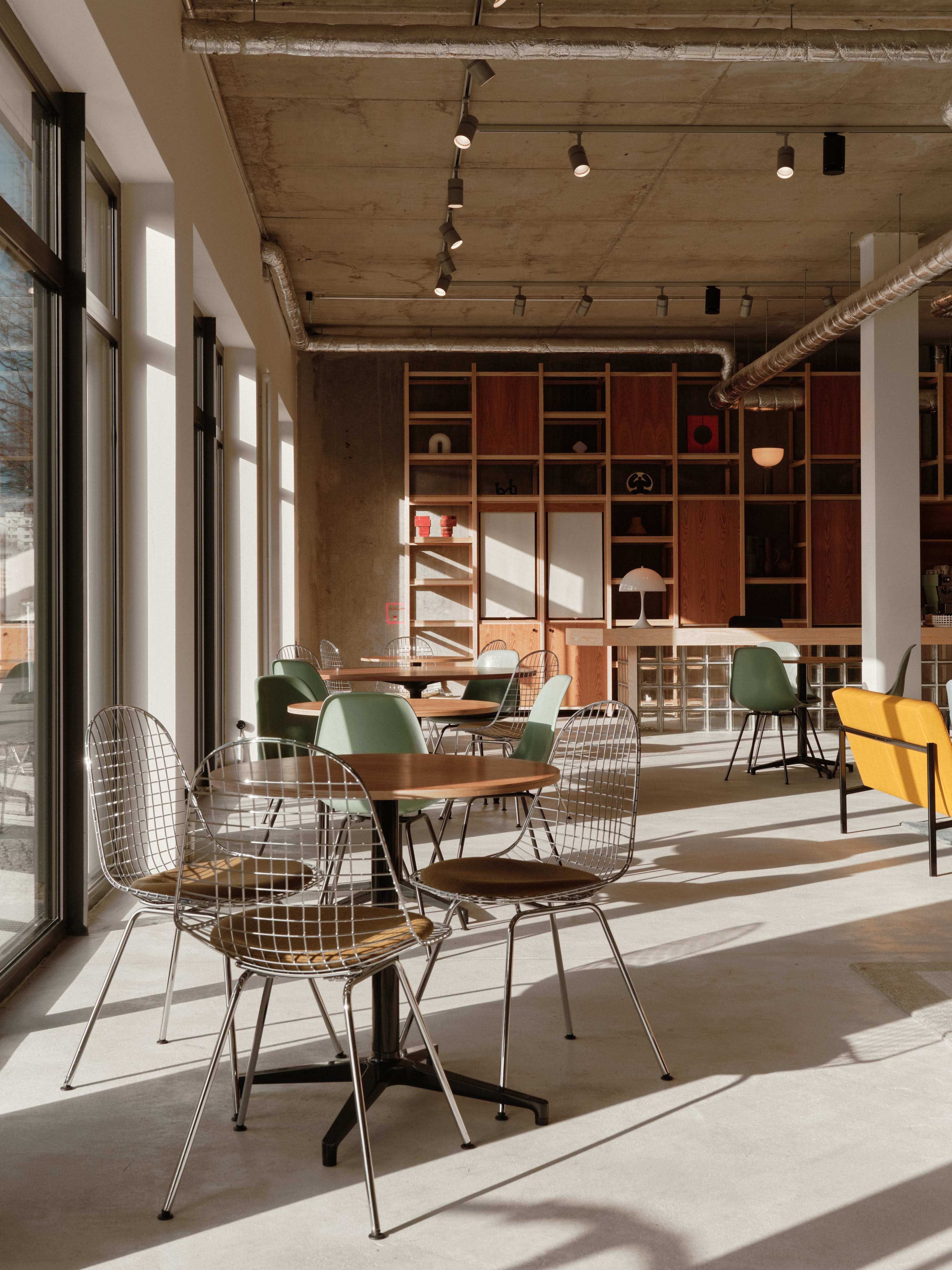 The image shows a modern café interior with various round tables and wire chairs, featuring a mix of colorful seating and a wooden shelving unit in the background.
