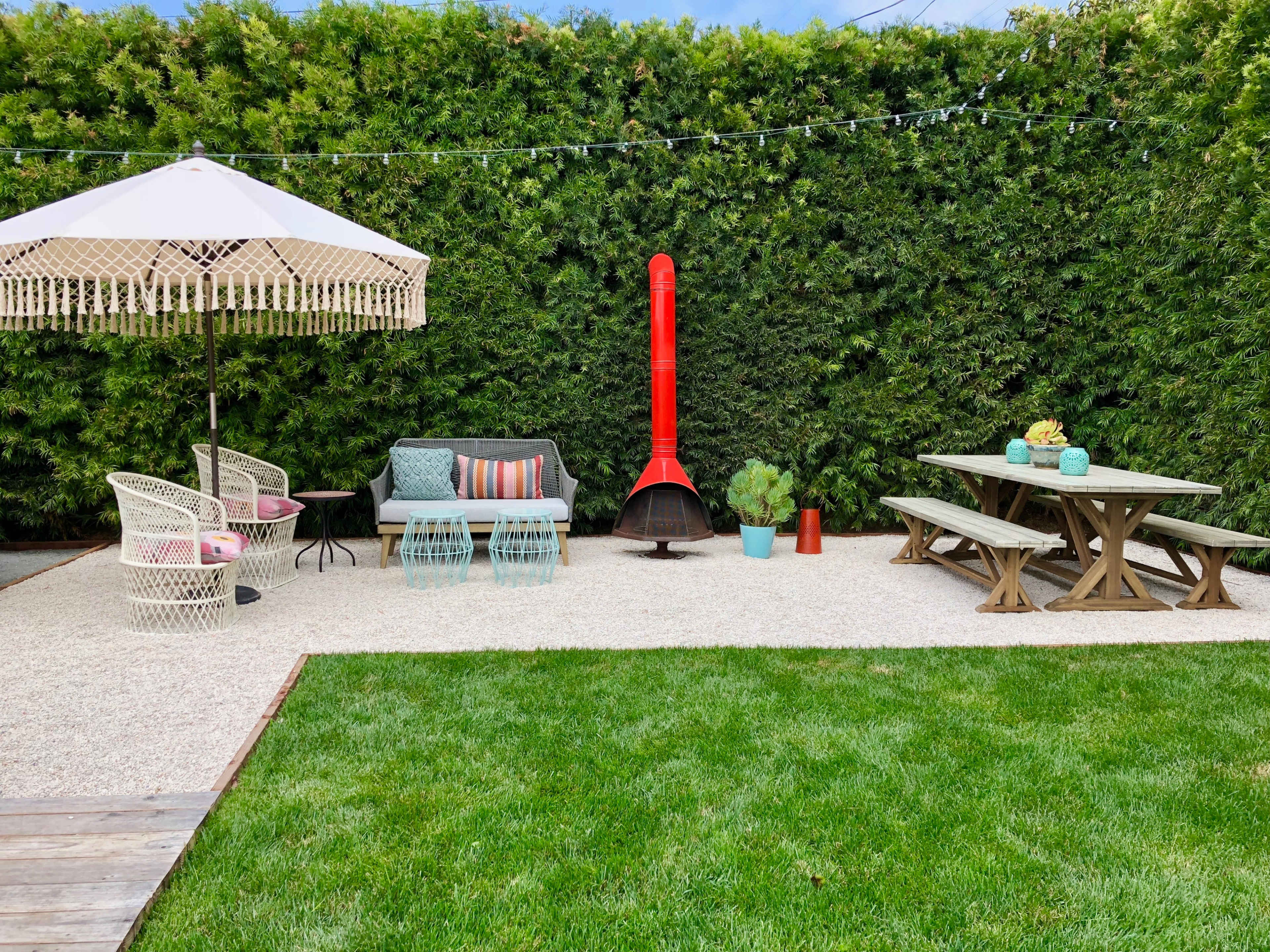The outdoor space features a vibrant seating area with a red outdoor fireplace, surrounded by lush green hedges and a gravel patio.