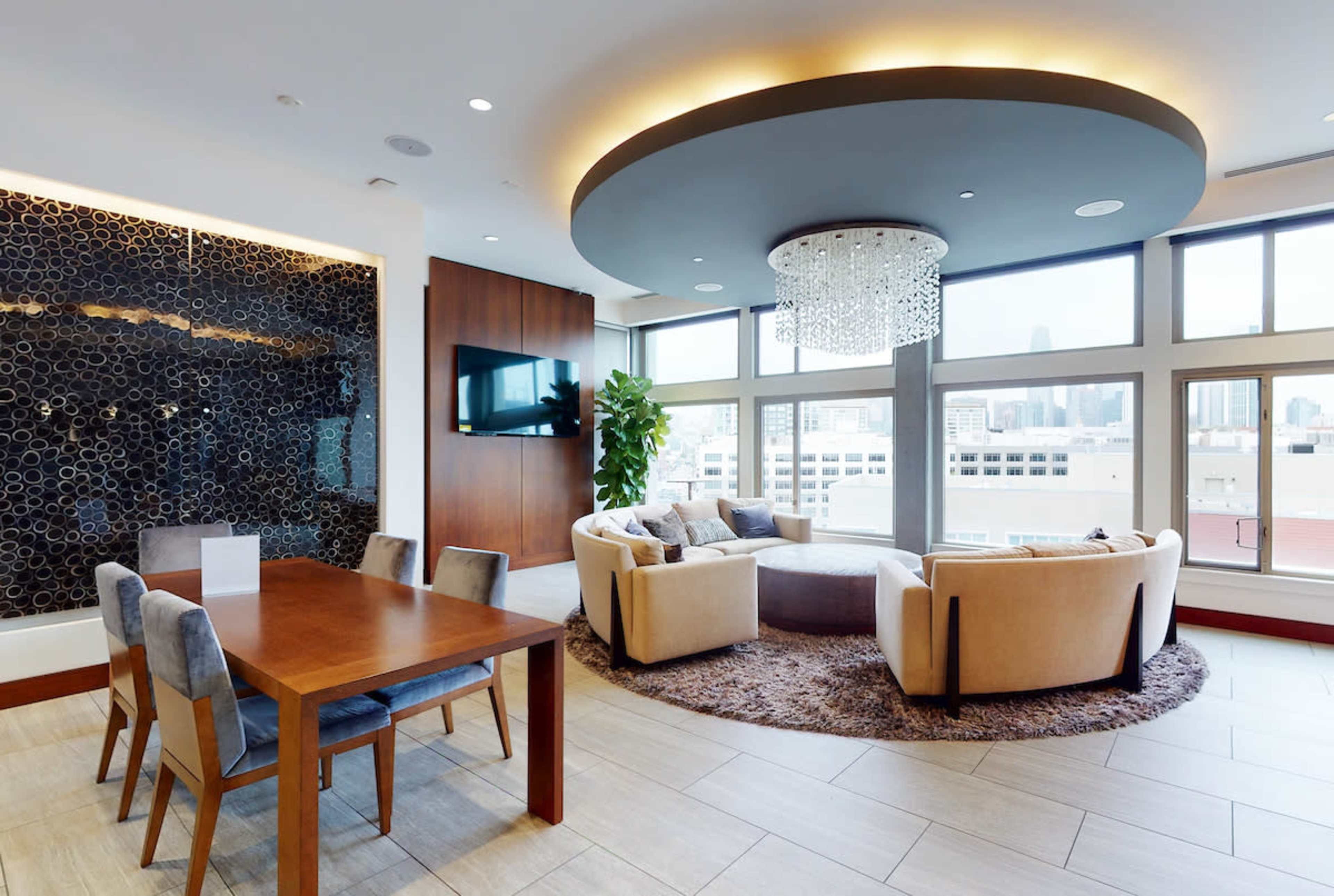 A modern living area features a round seating arrangement, dining table, and large windows with city views.