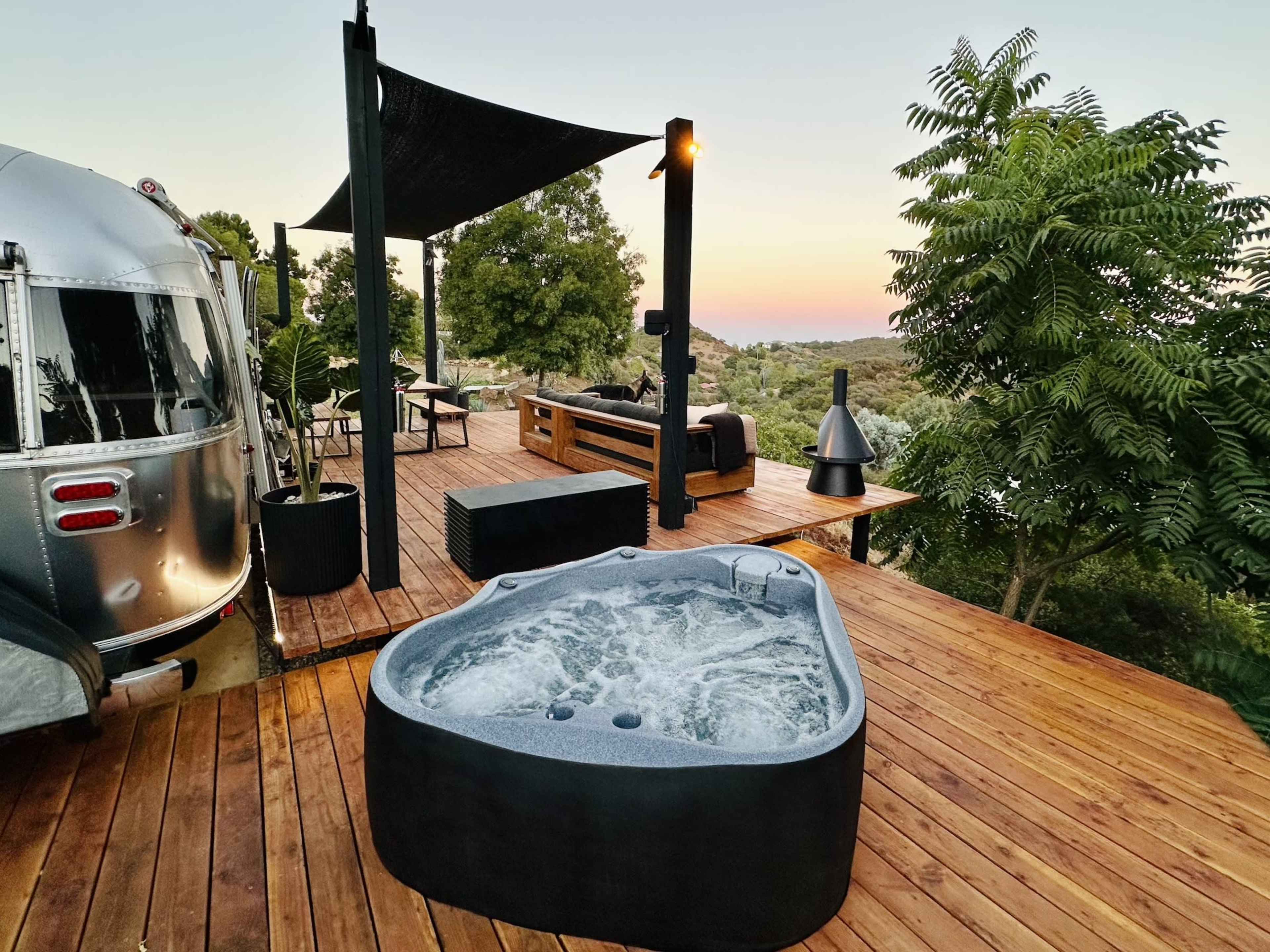 A modern hot tub sits on a wooden deck beside a parked Airstream trailer, with outdoor seating and greenery in the background.