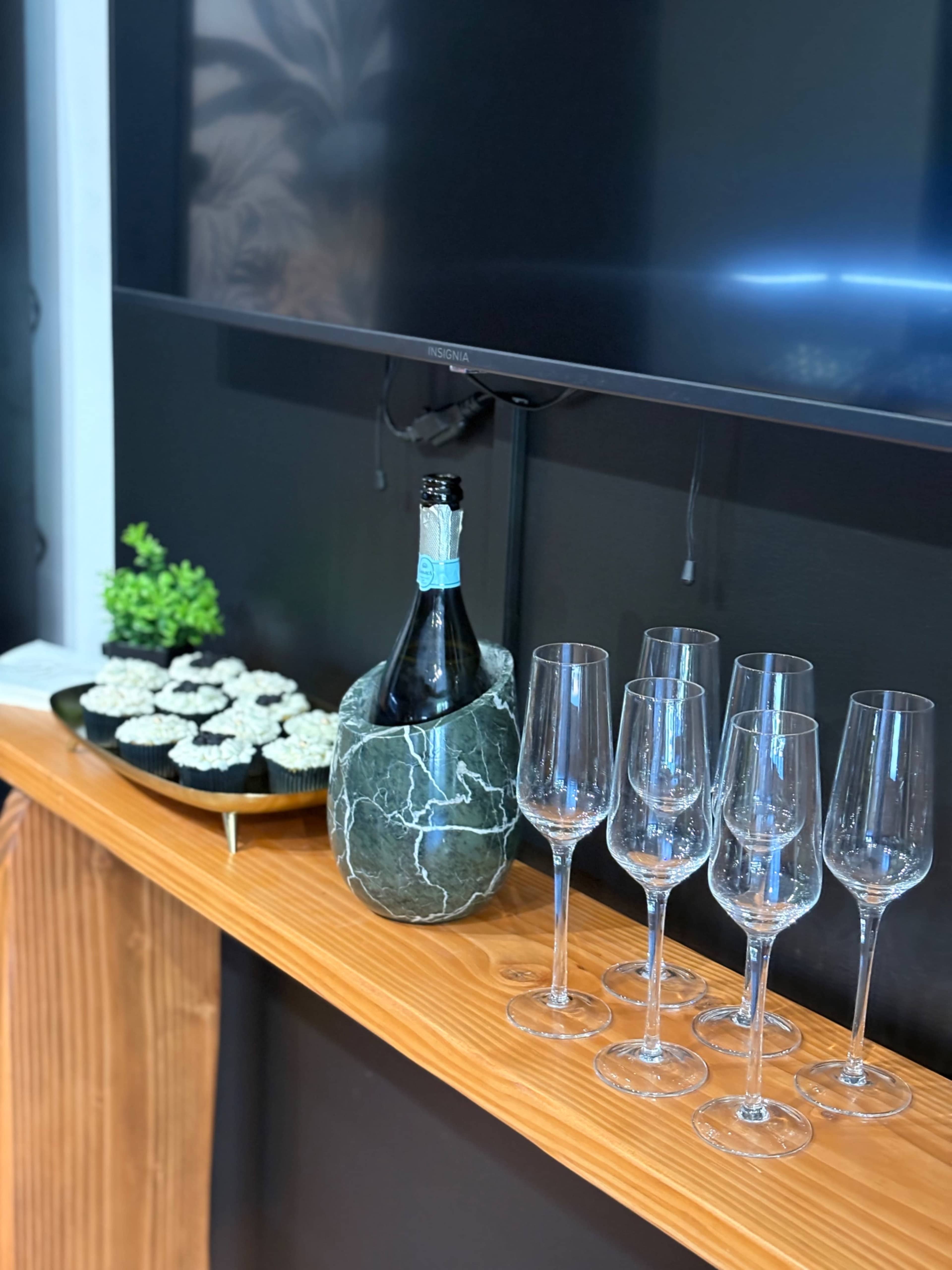 A bottle of sparkling wine is placed in a stone cooler beside a black tray of sushi and several clear champagne glasses on a wooden shelf.