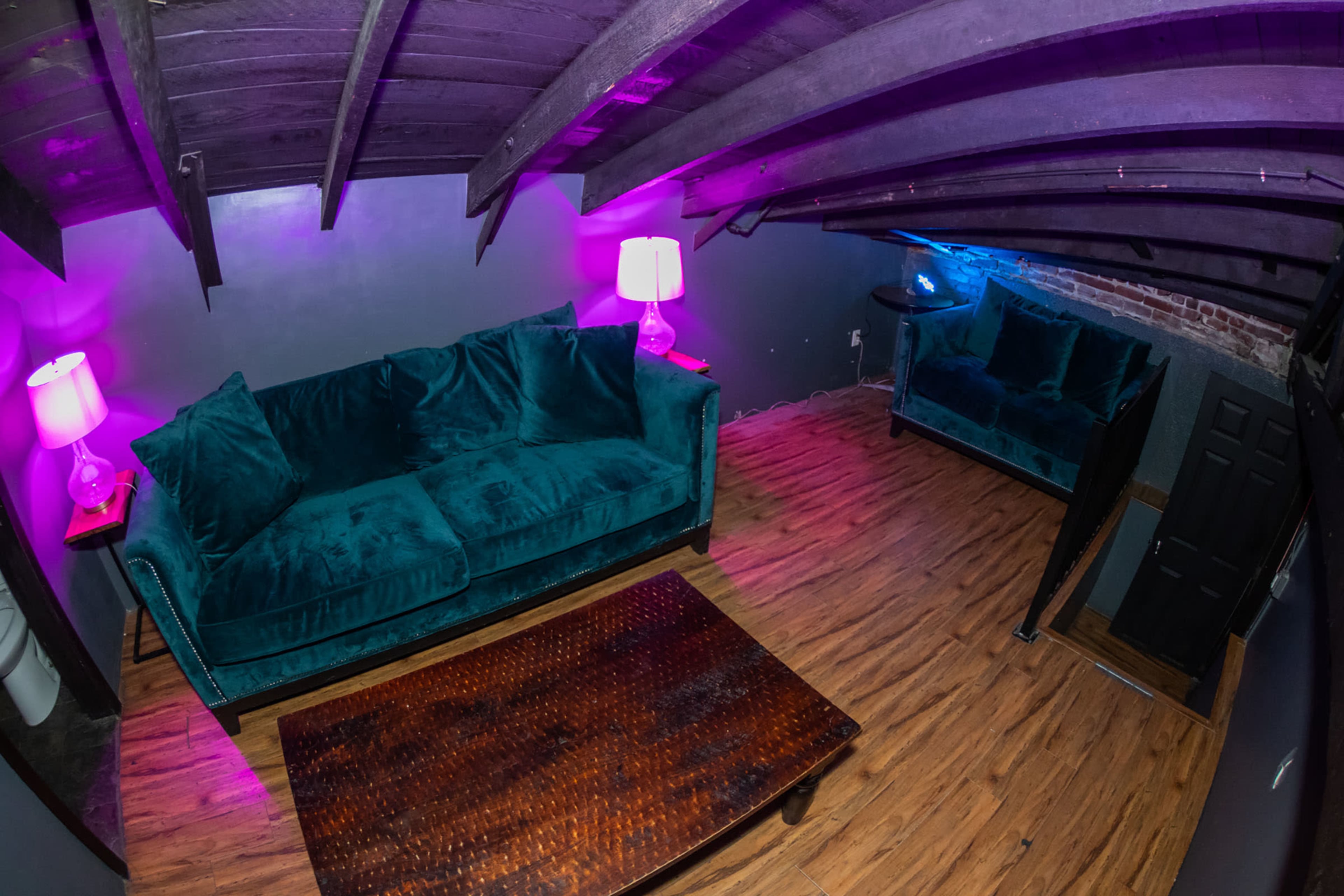 A cozy living space features a green velvet sofa, a wooden coffee table, and lamps emitting purple and blue light, all under a beamed ceiling.