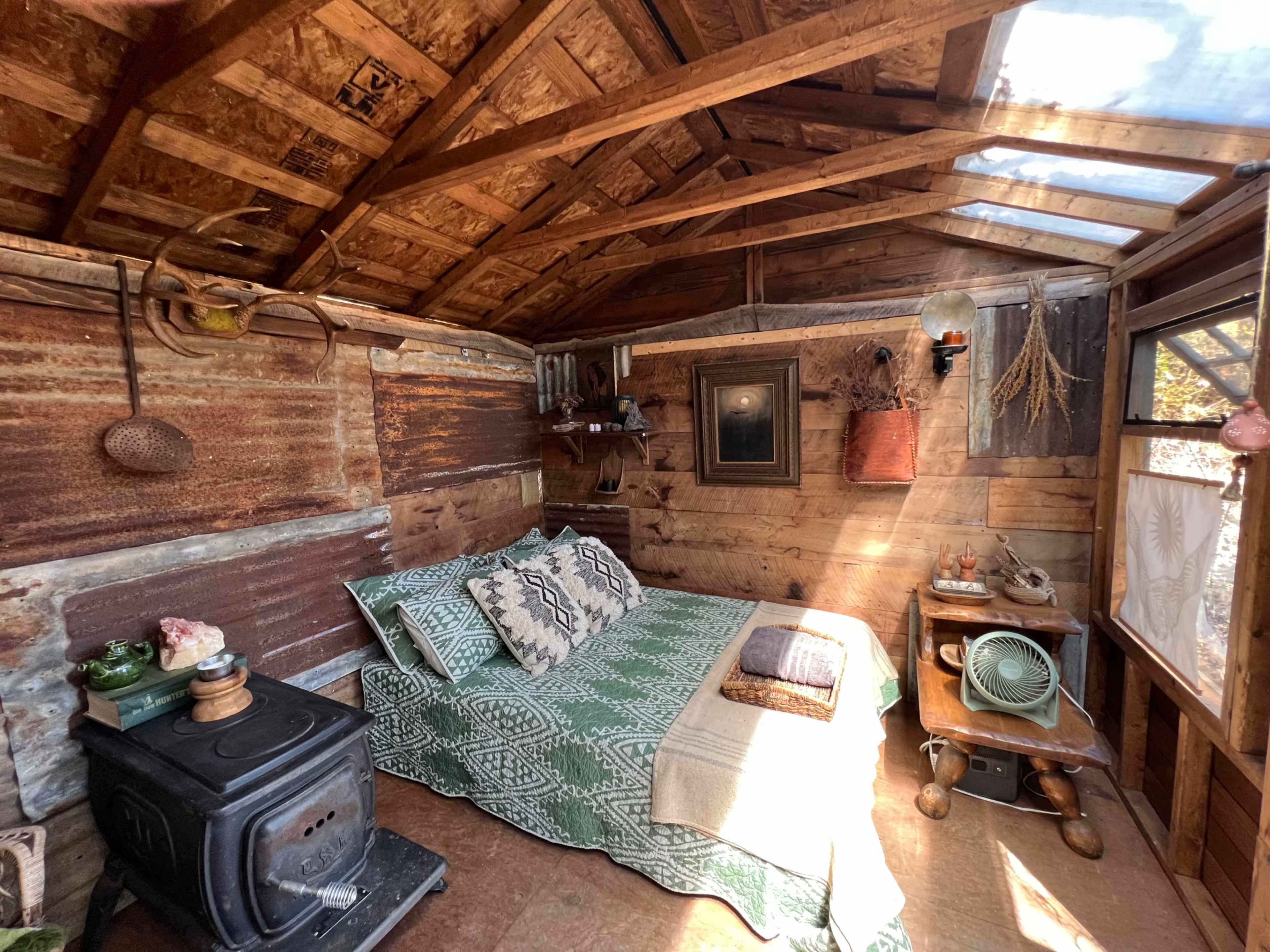 The interior of a rustic cabin features a bed with a patterned blanket, a wood stove, and various decorations on the walls and furniture.