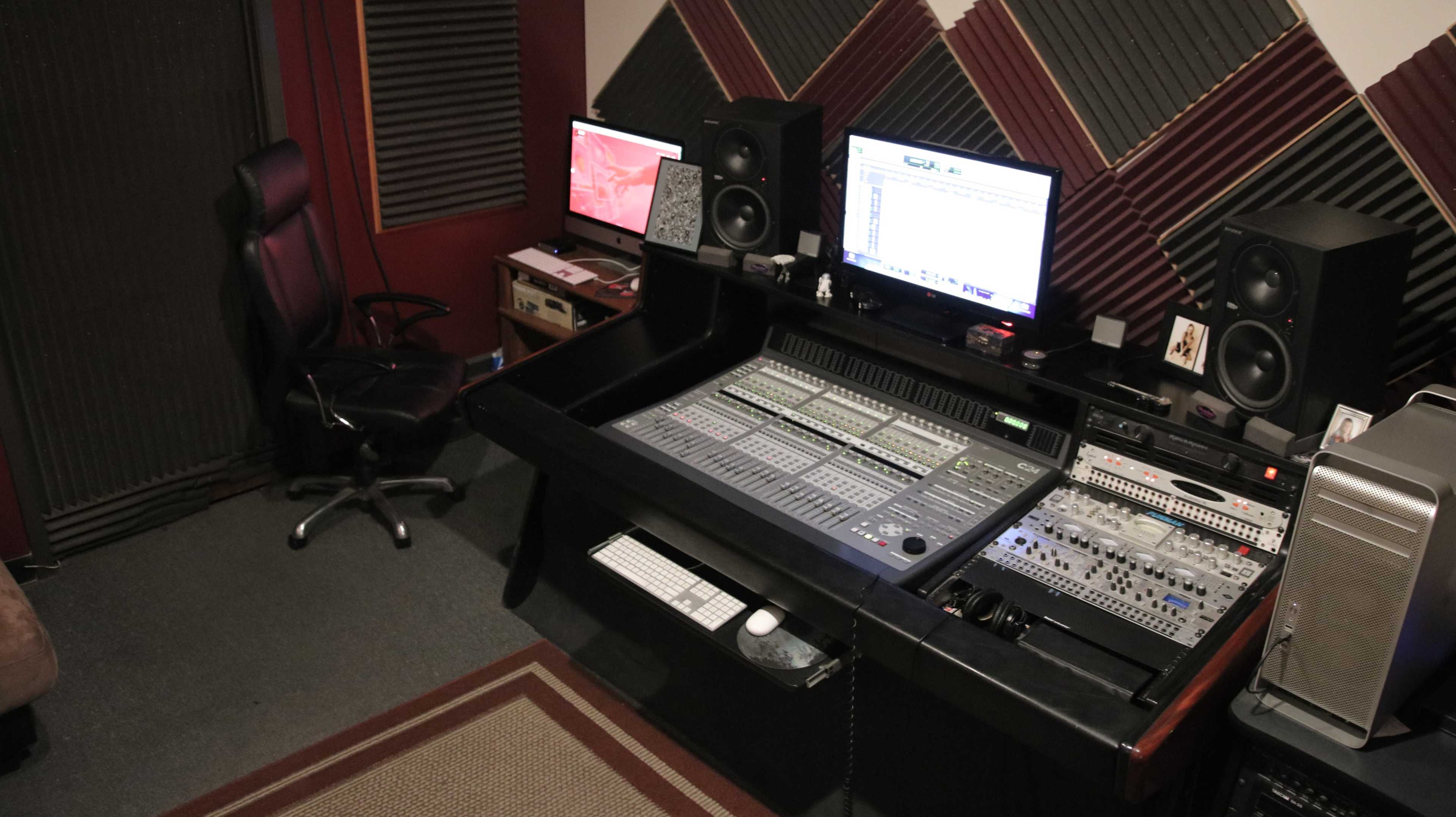 A music production studio features a large mixing console, dual computer monitors, studio speakers, and a black office chair in a soundproofed room.