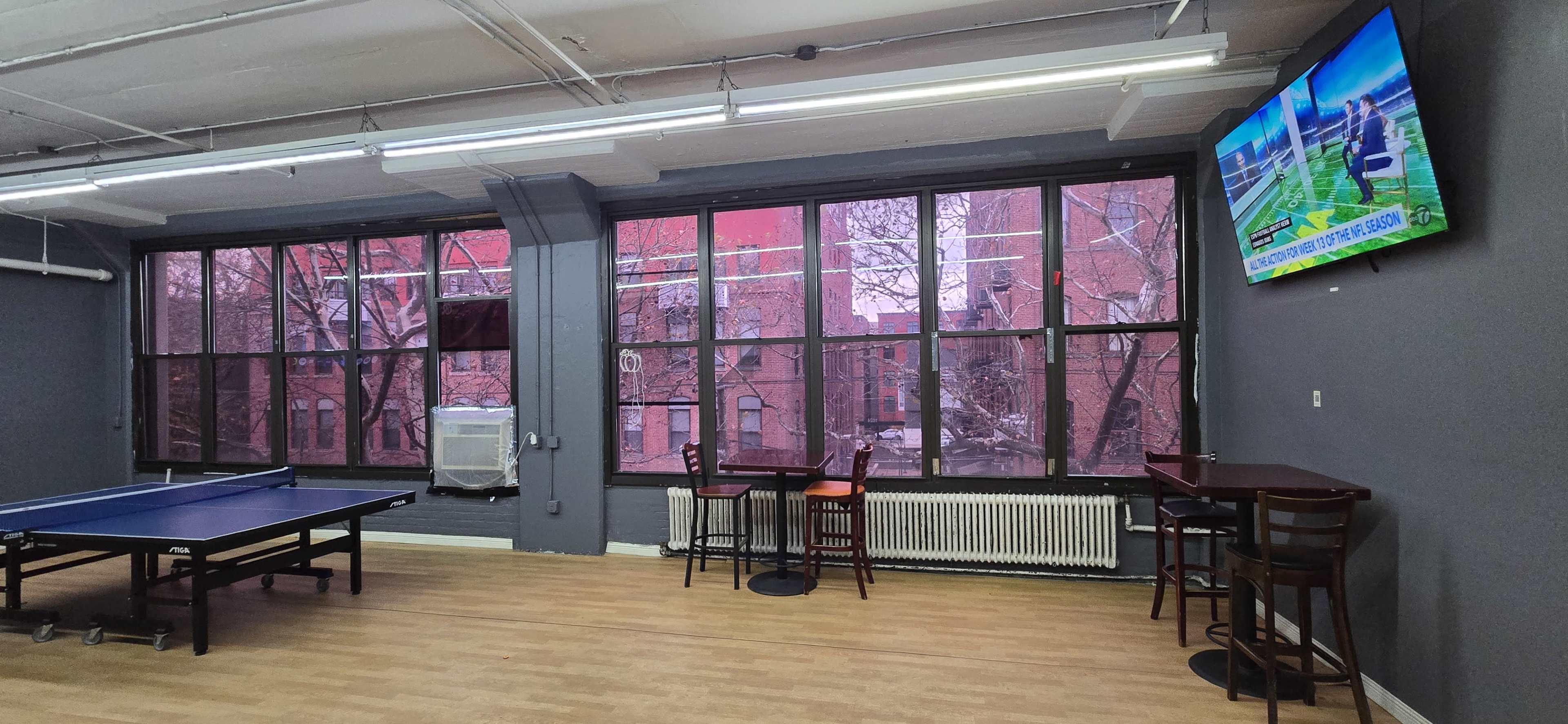 The image shows a recreational room with a ping pong table, bar-style seating, large windows displaying a pink-hued exterior, and a wall-mounted television.