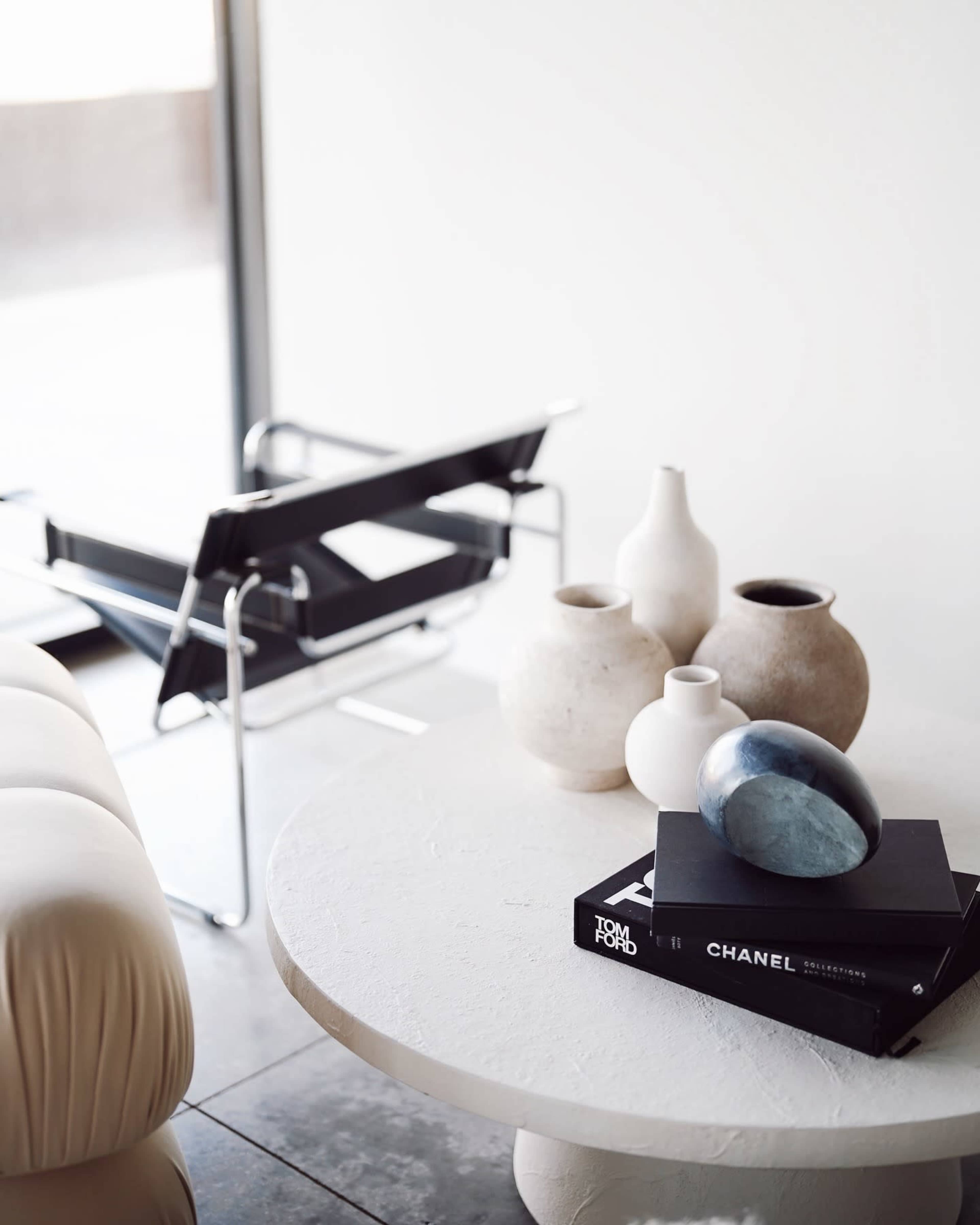A modern living space features a round table with decorative stones and books, surrounded by minimalist vases and a black chair.