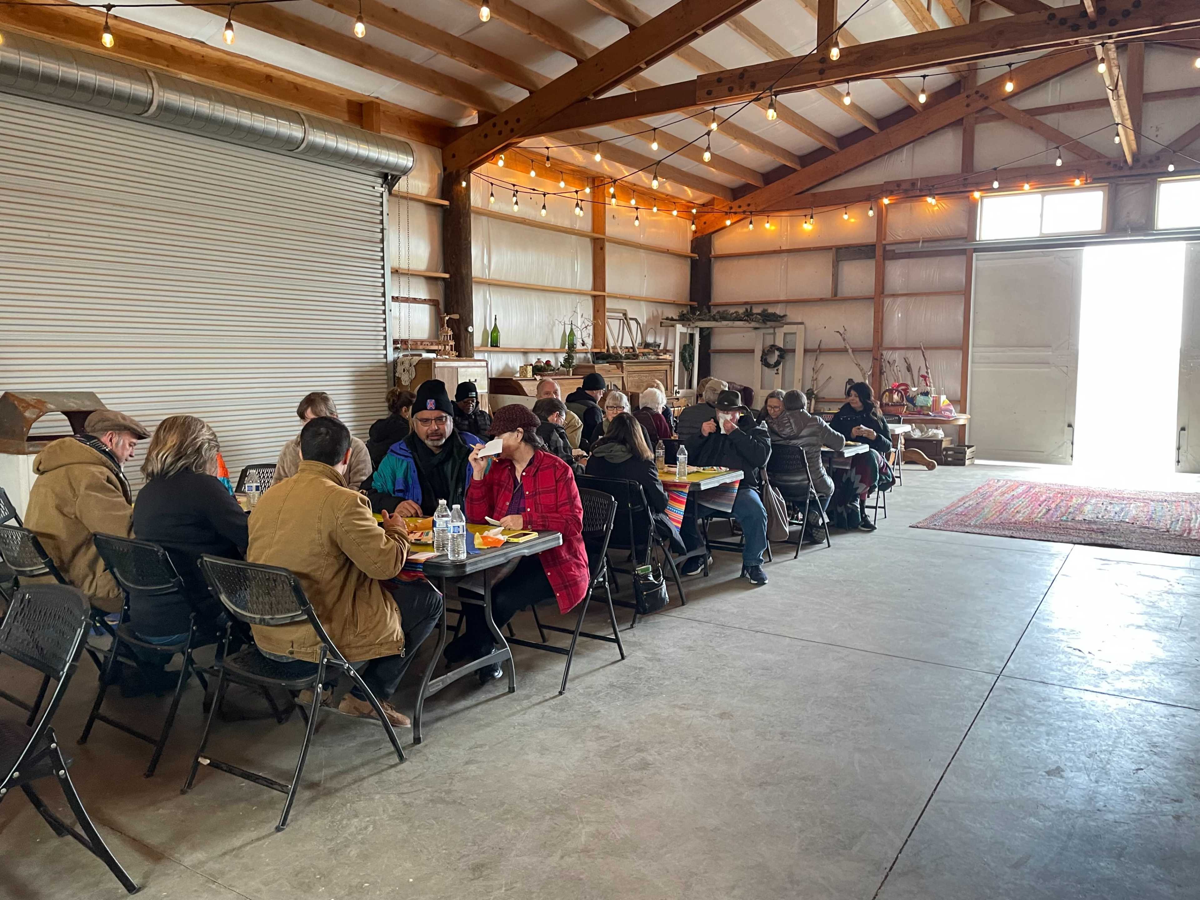 A group of people sits at tables in a spacious, well-lit barn, sharing a meal together.