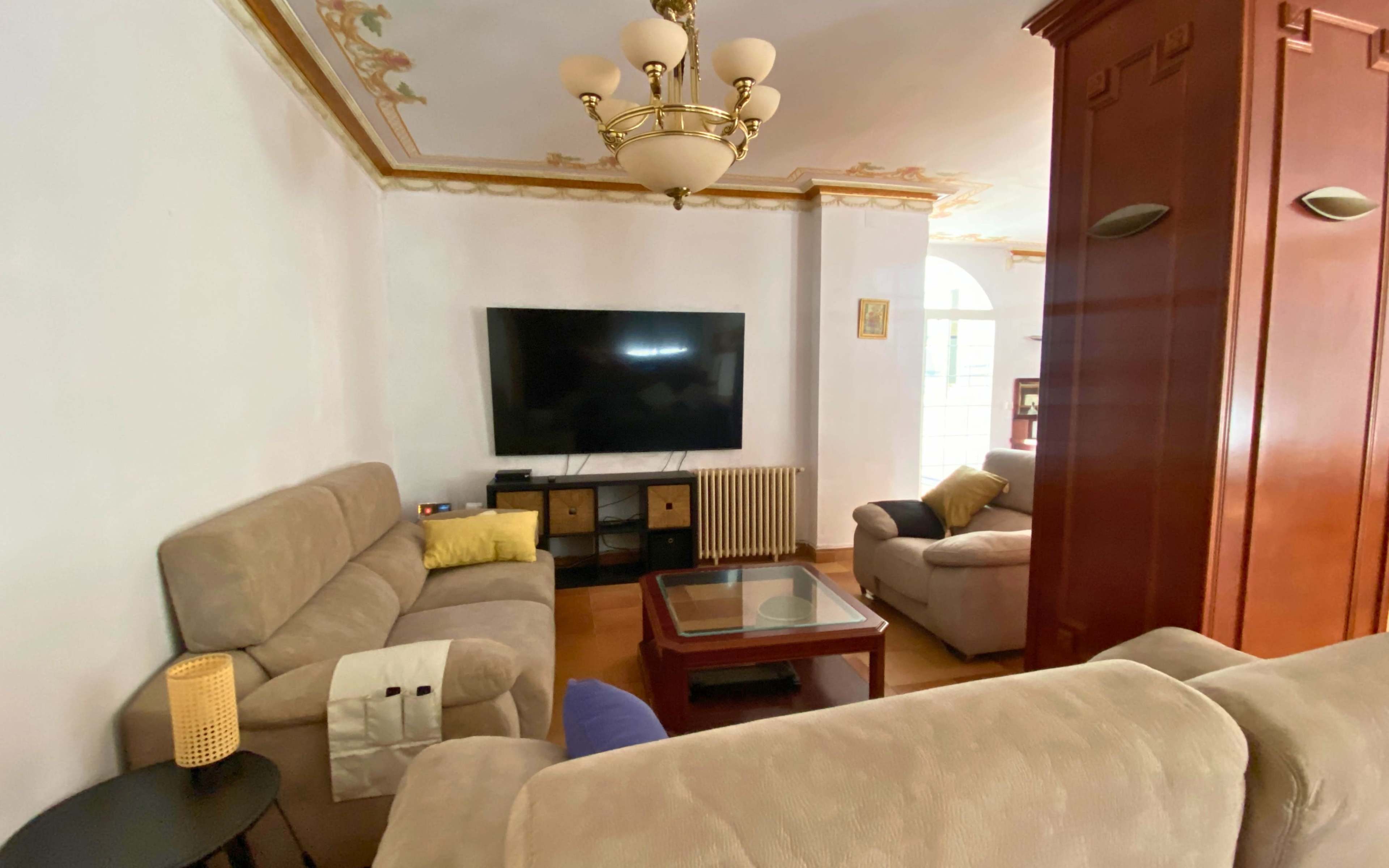 The image shows a cozy living room with two couches, a coffee table, a flat-screen TV mounted on the wall, and wooden furniture in the background.