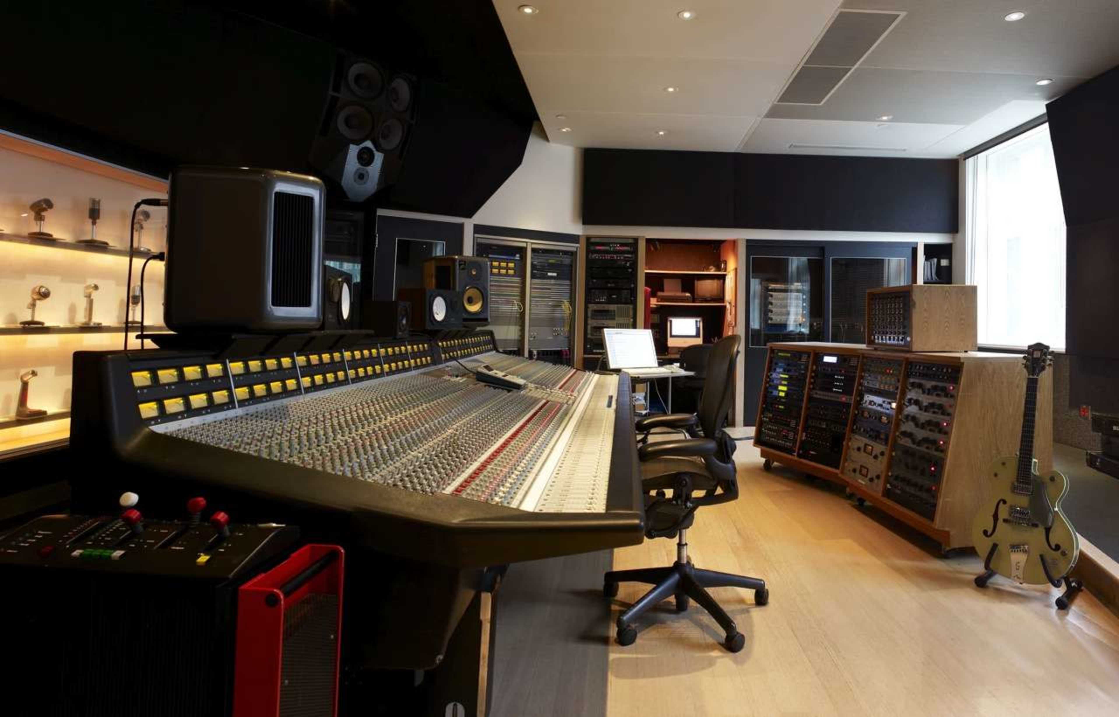 The image shows a recording studio with a large mixing console, several audio equipment racks, and a guitar displayed in the corner.