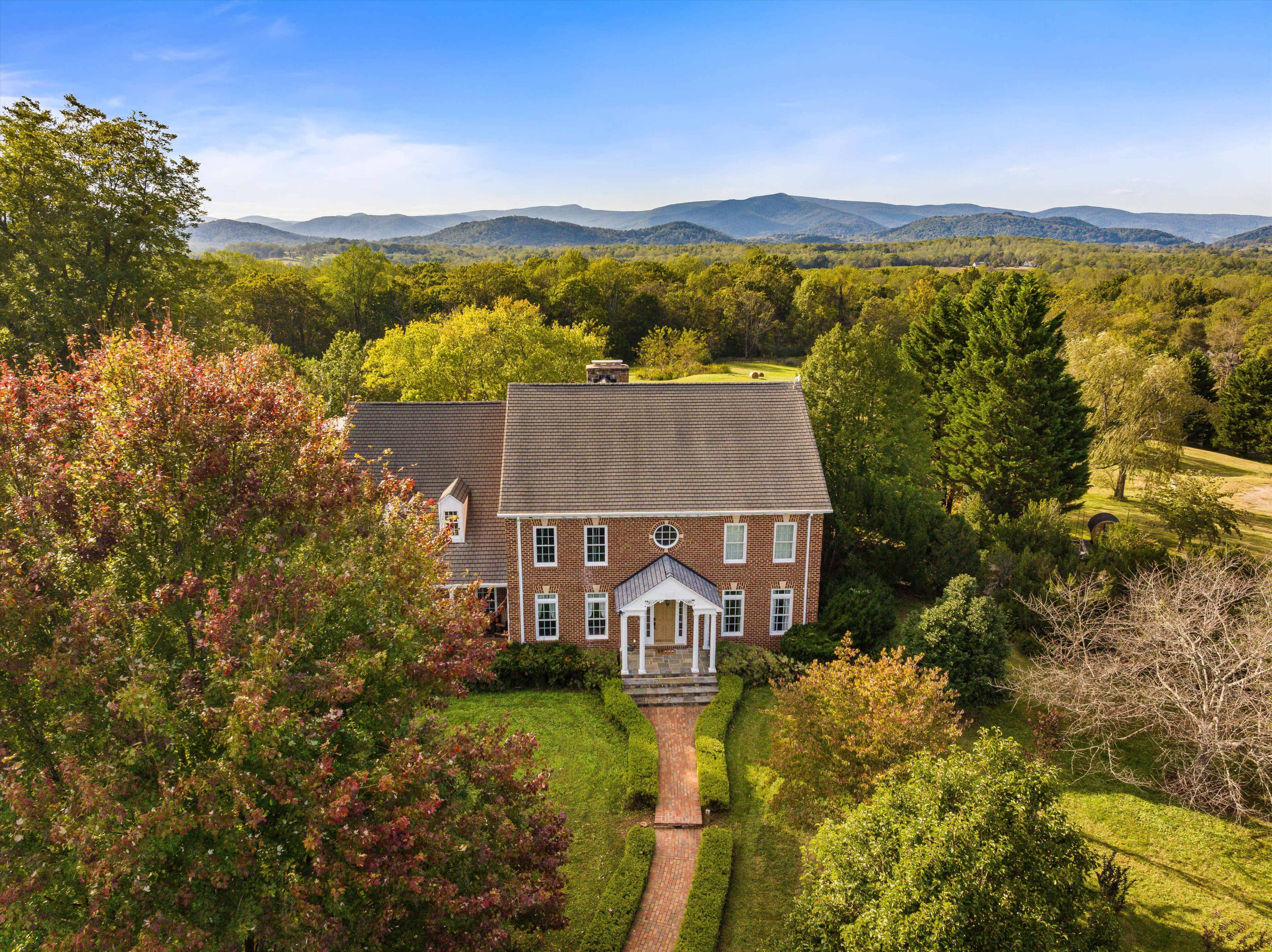 A large brick house with a well-manicured lawn and a winding pathway leads to the front entrance, surrounded by trees and mountains in the background.