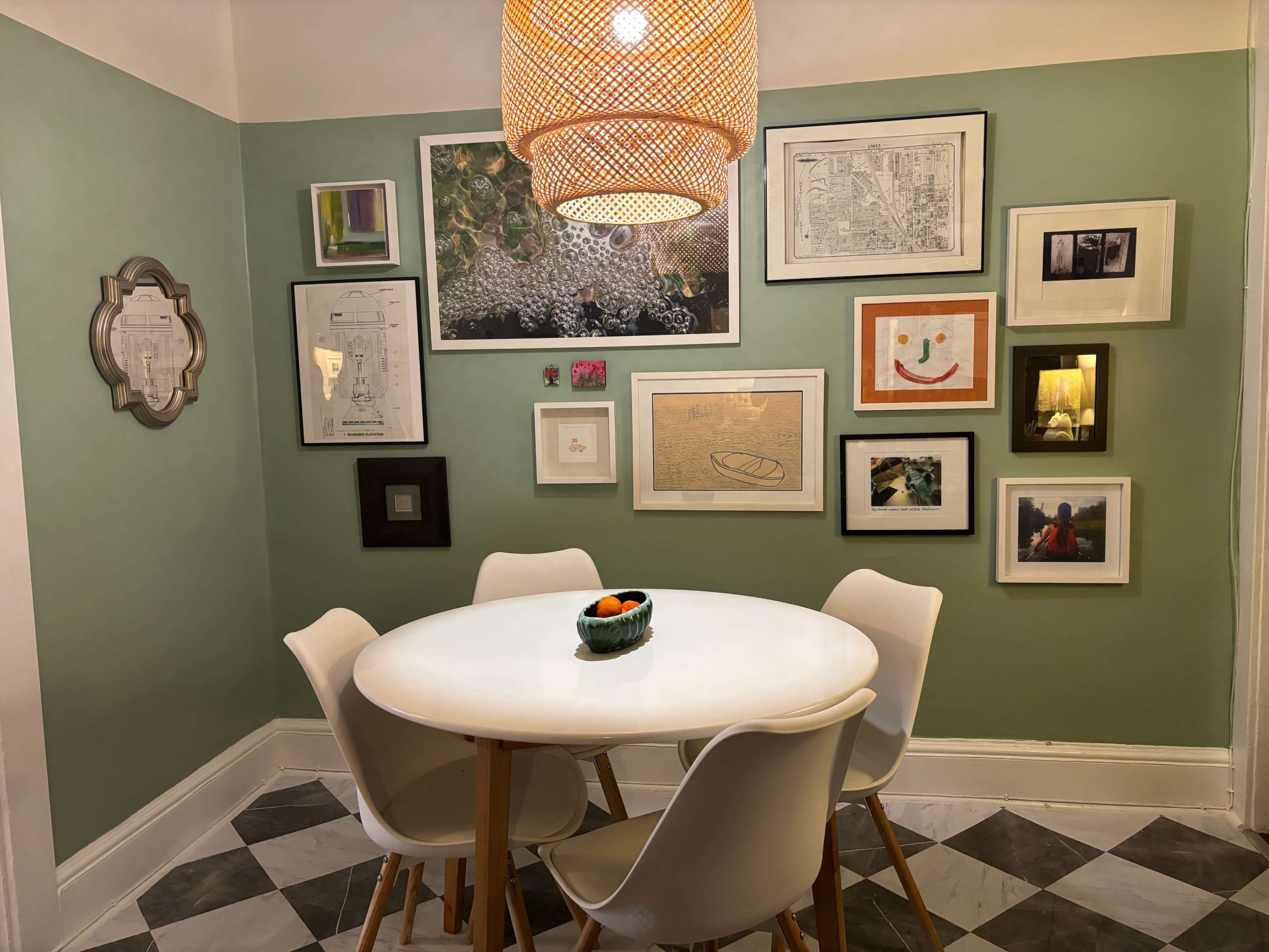A round white table with four chairs is positioned in a corner of a room adorned with various framed artworks on green walls.