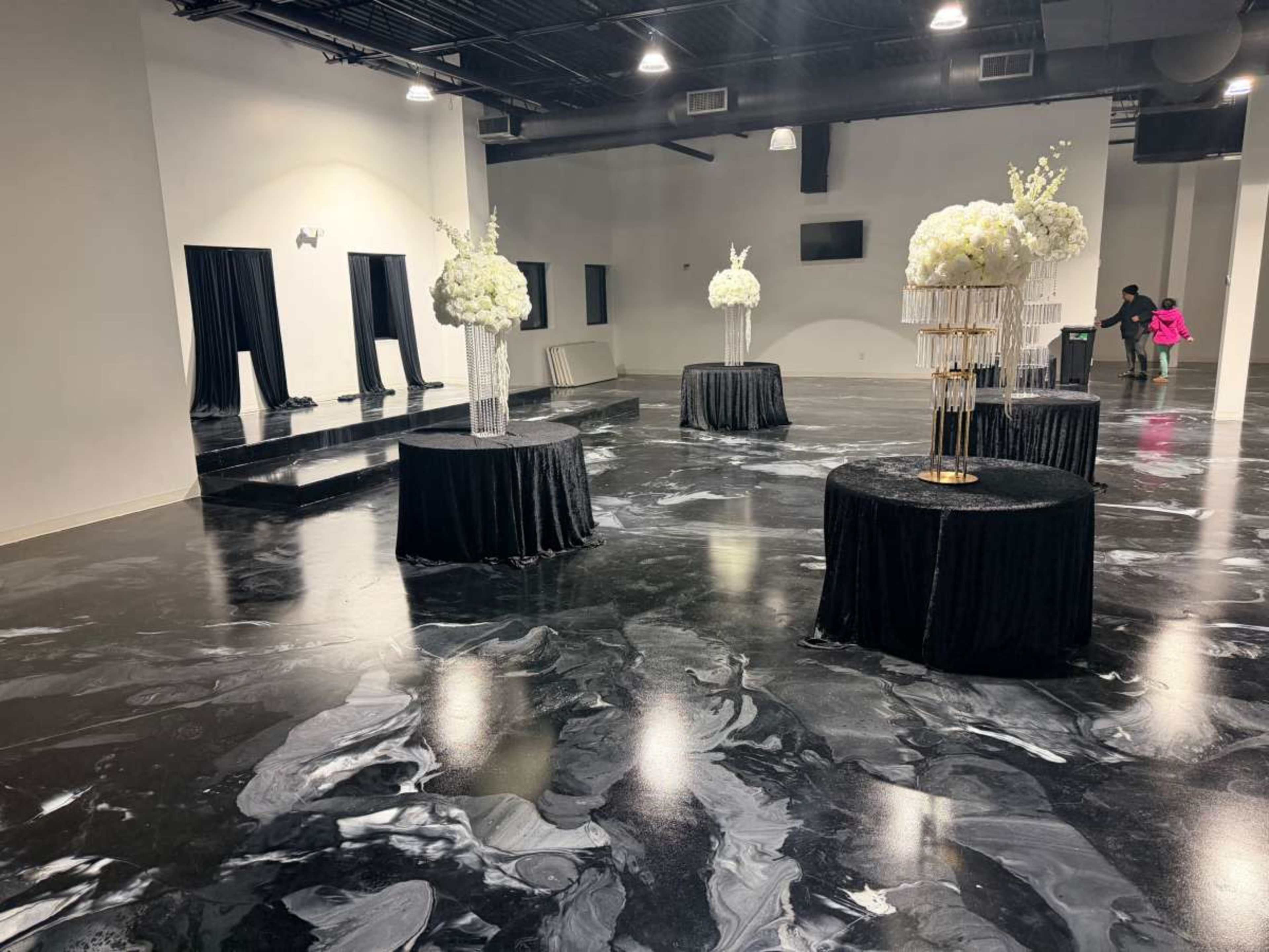 The image shows a spacious event room with a dark, marbled floor, featuring five circular tables adorned with black tablecloths and floral centerpieces.