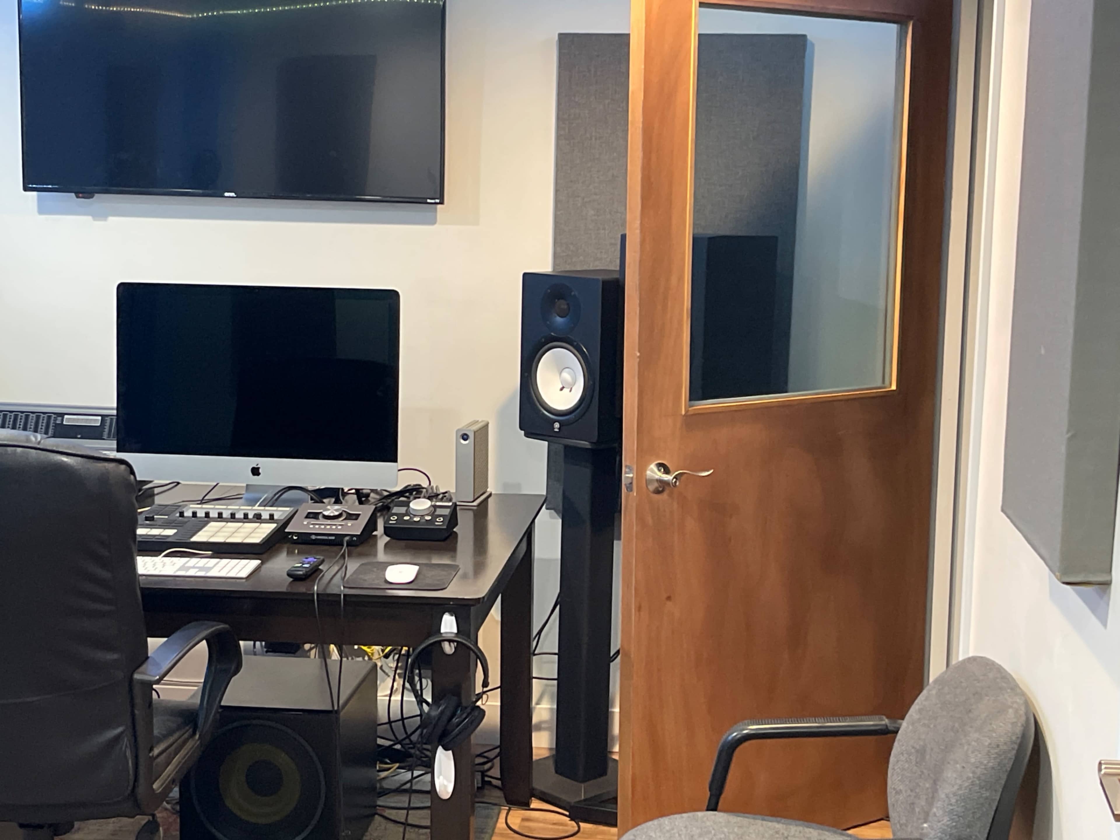 The image shows a music studio with a desk featuring a computer, speakers, and audio equipment, next to a door leading to another room.
