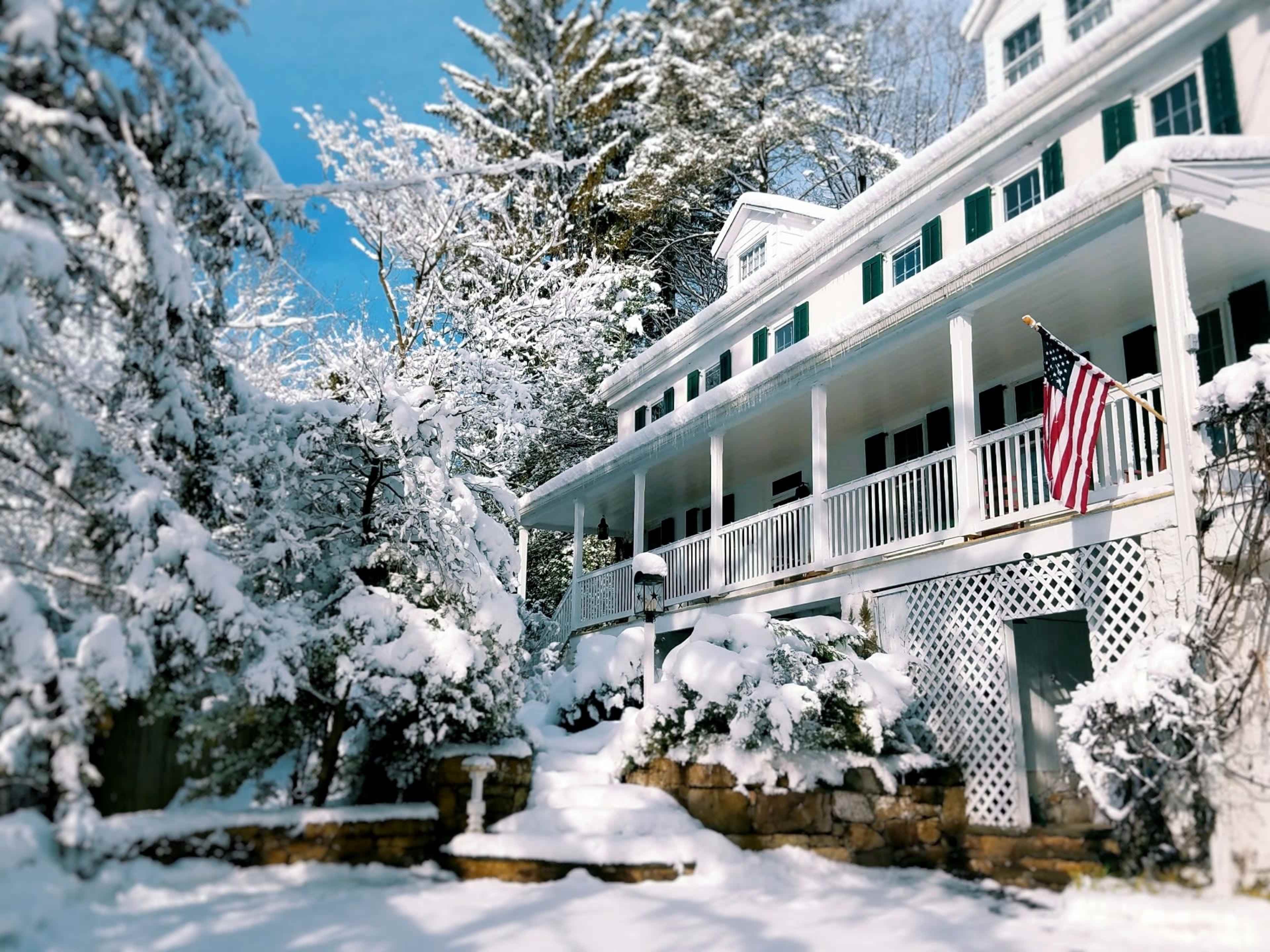 A large white house with a covered porch is surrounded by snow-covered trees, and an American flag hangs from the balcony.