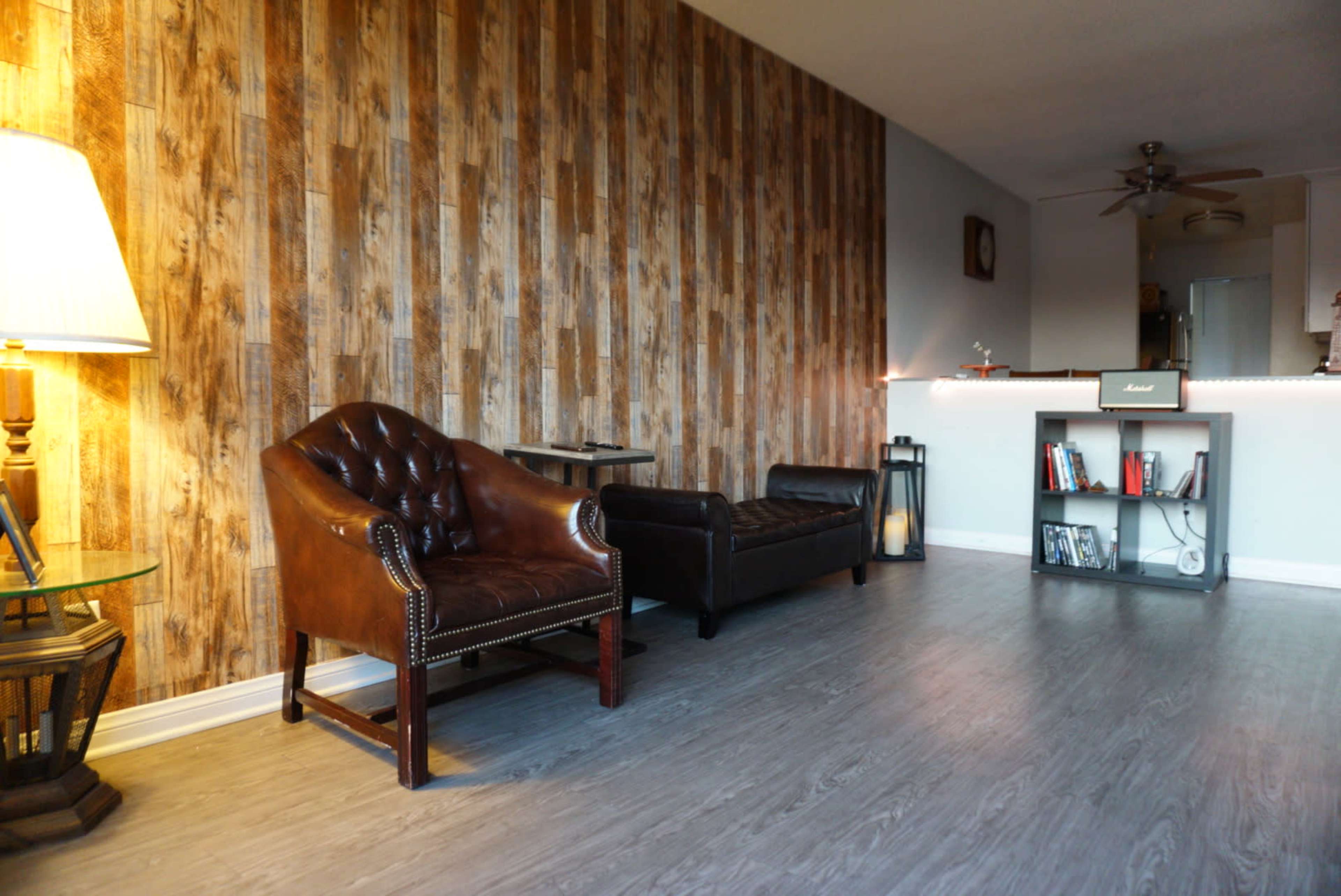 A living room with a wood-paneled wall, a brown leather armchair, a dark leather sofa, a glass side table with a lamp, and a shelf with books and a small speaker.