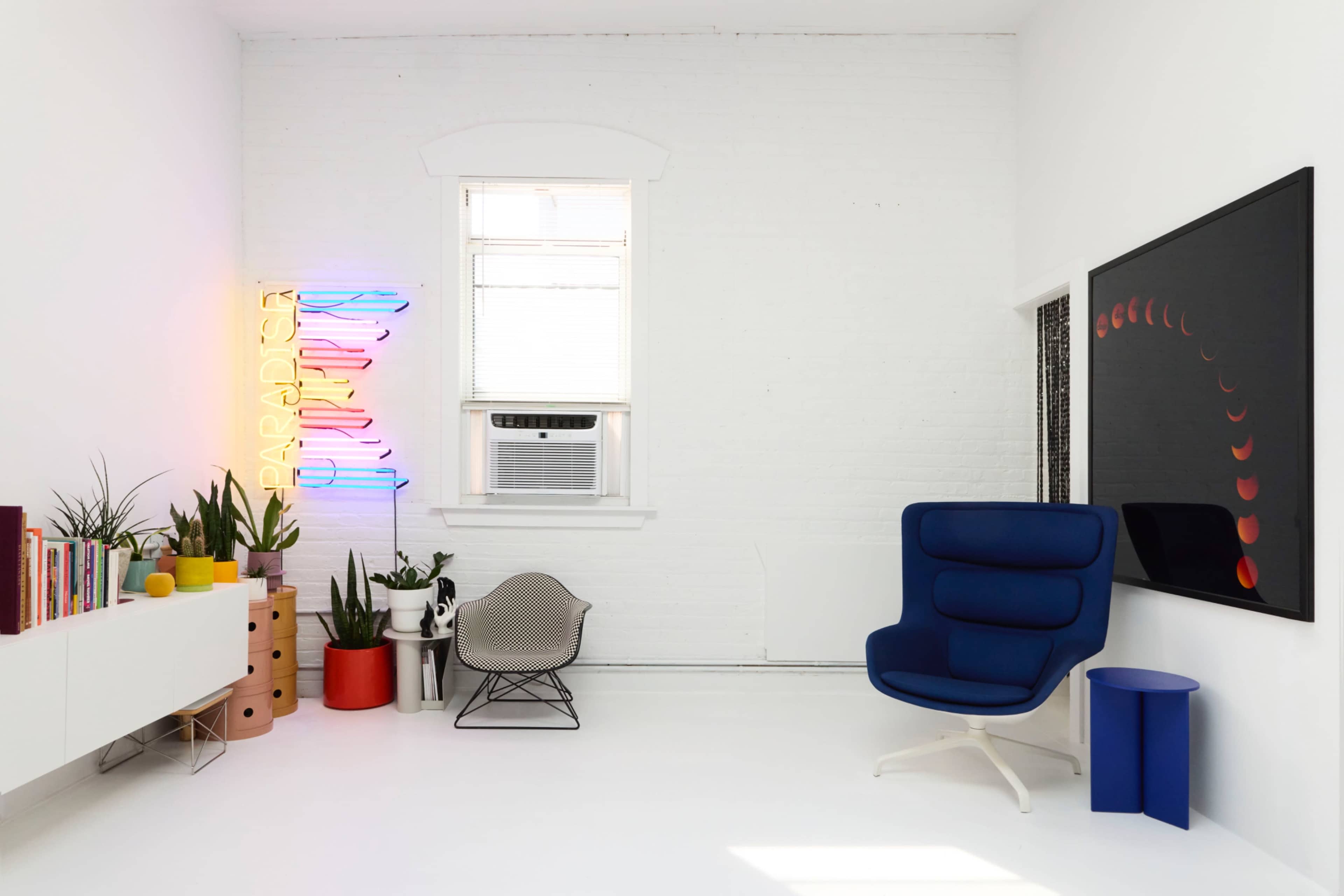 The image shows a modern, light-filled room featuring a blue chair, a colorful neon sign, and various indoor plants arranged around a white furniture piece.