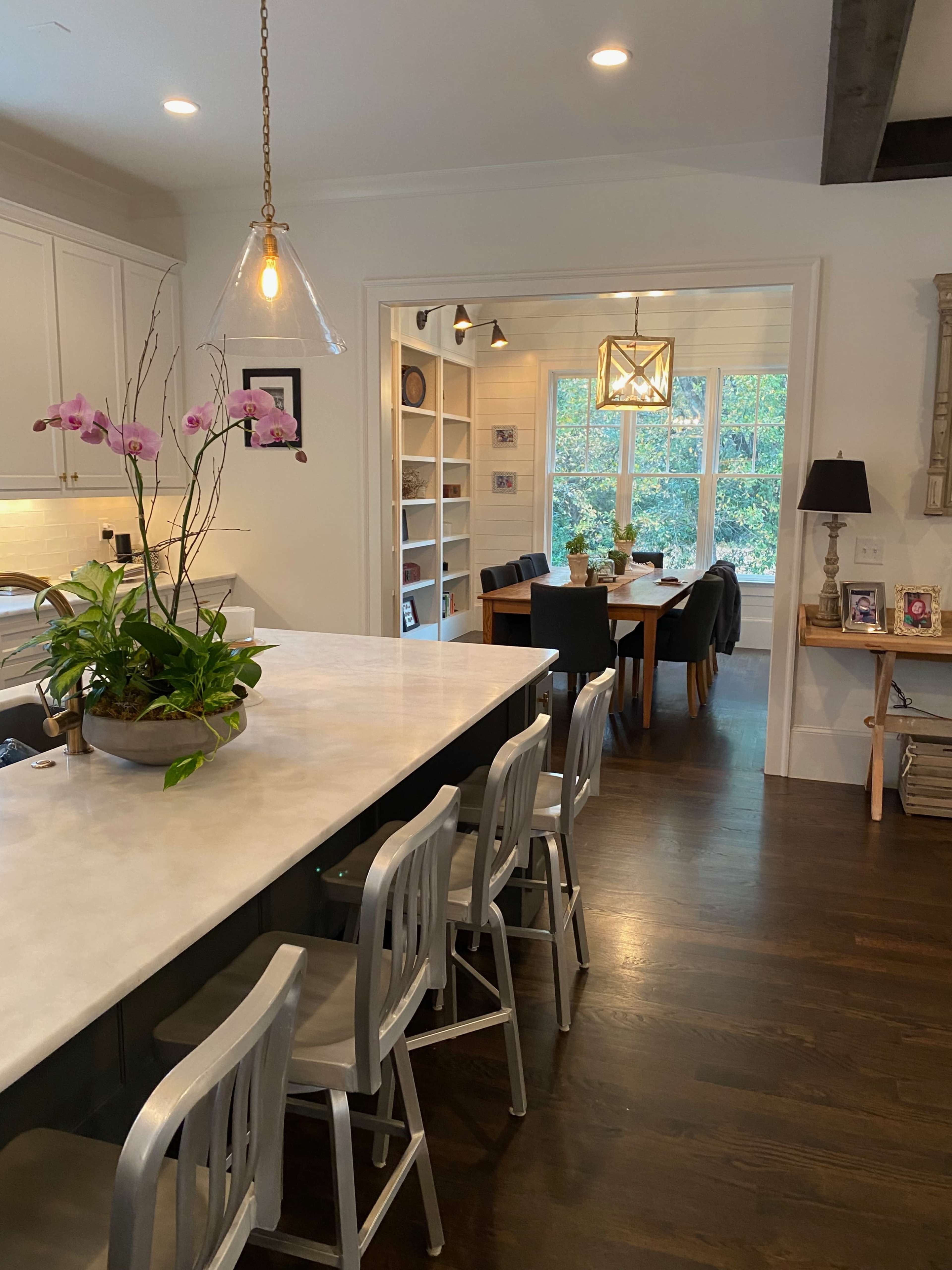 The image shows a kitchen with a marble island and bar stools, leading into a dining area with a table surrounded by chairs and large windows.
