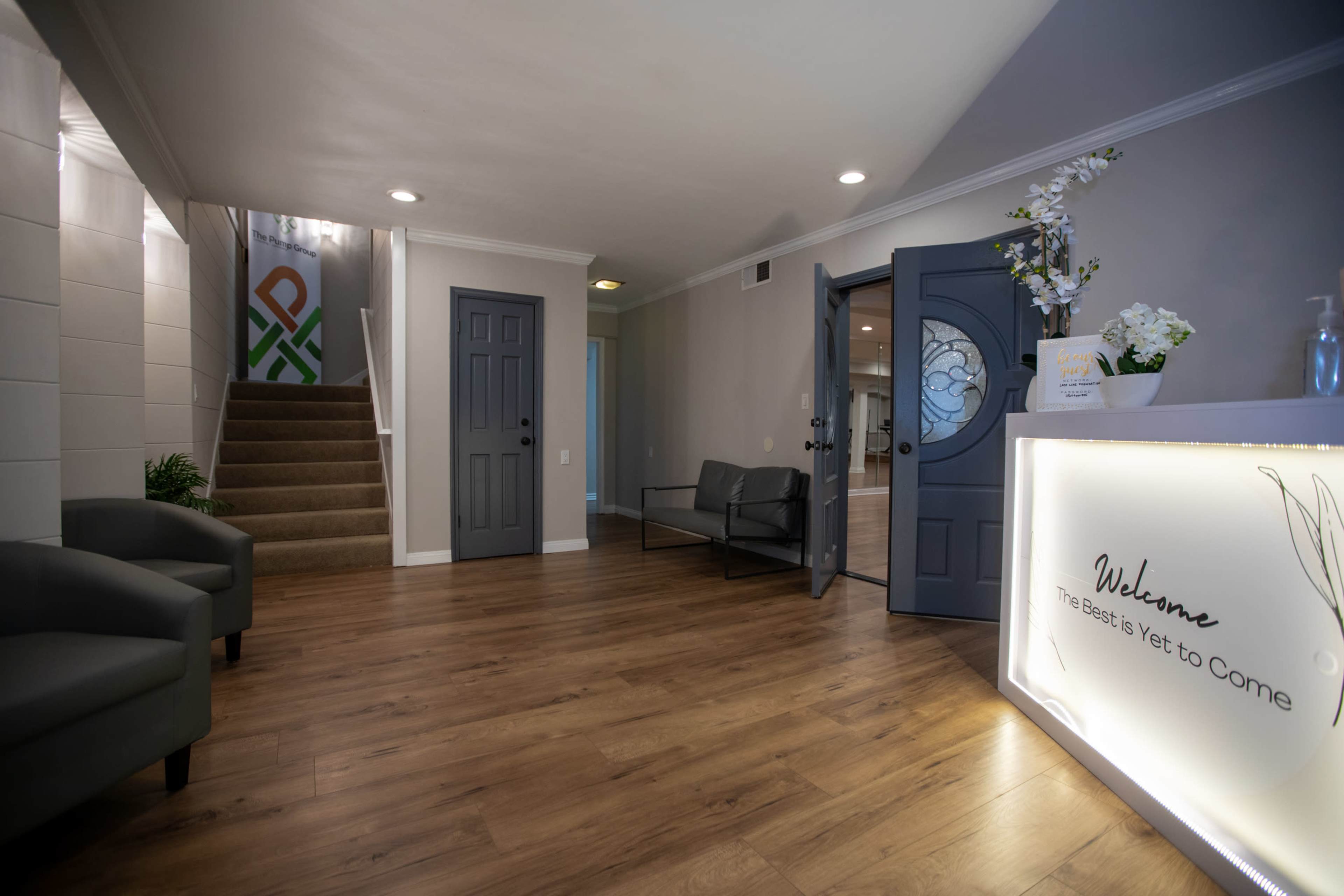 The image shows a welcoming reception area with a sleek modern design, featuring a lit reception desk, seating areas, and a staircase leading to an upper level.
