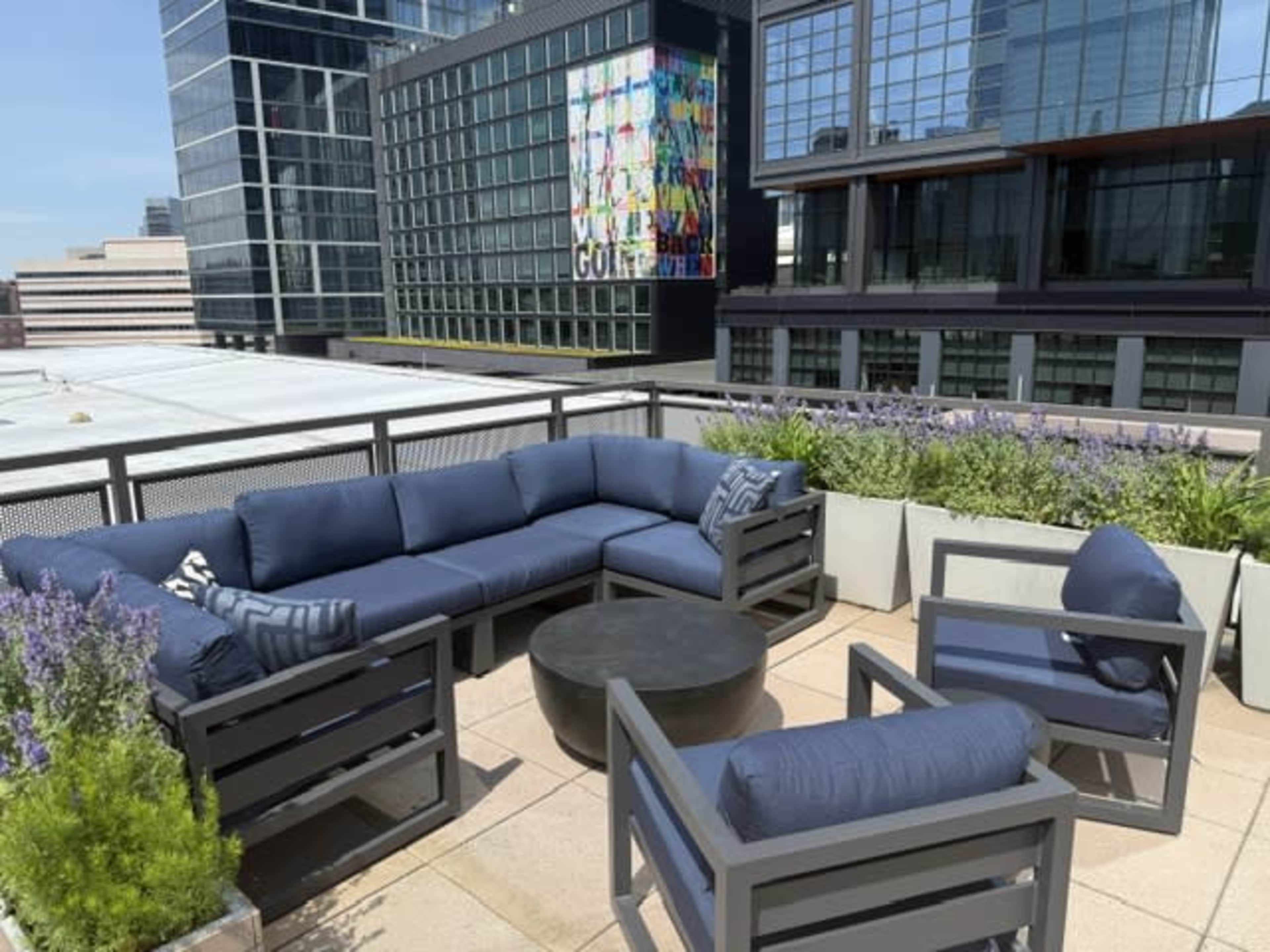 The image shows a rooftop patio with modern seating arrangements, a round table, and a colorful mural on an adjacent building.