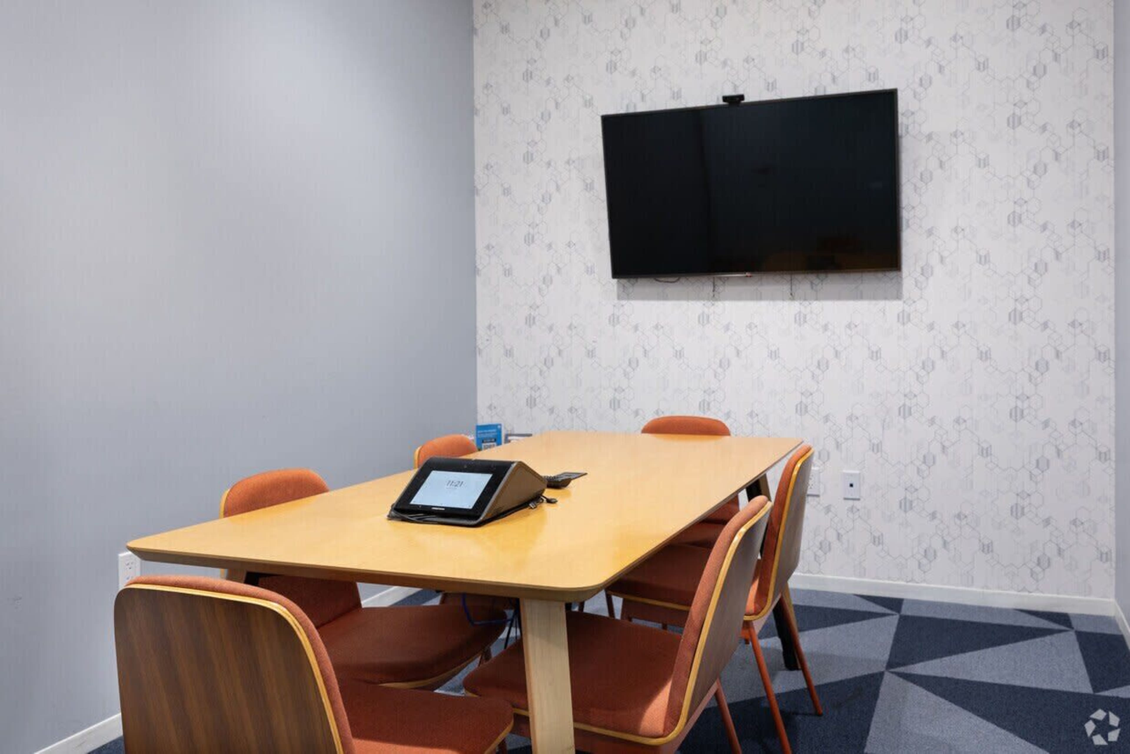 A conference room contains a wooden table with six orange chairs and a wall-mounted television.