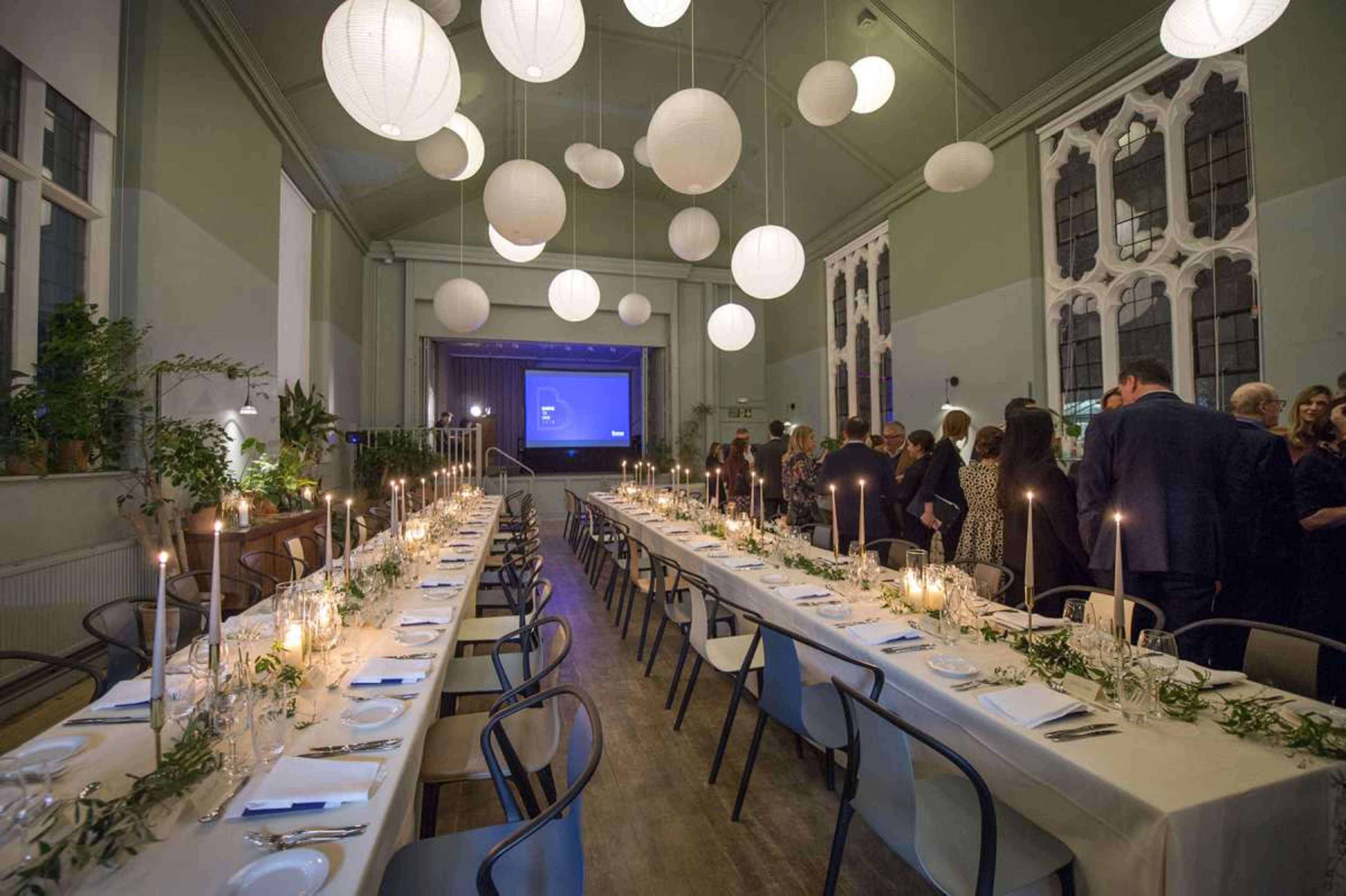 A long dining table is elegantly set with candles and greenery, surrounded by guests in a spacious venue with decorative paper lanterns hanging from the ceiling.