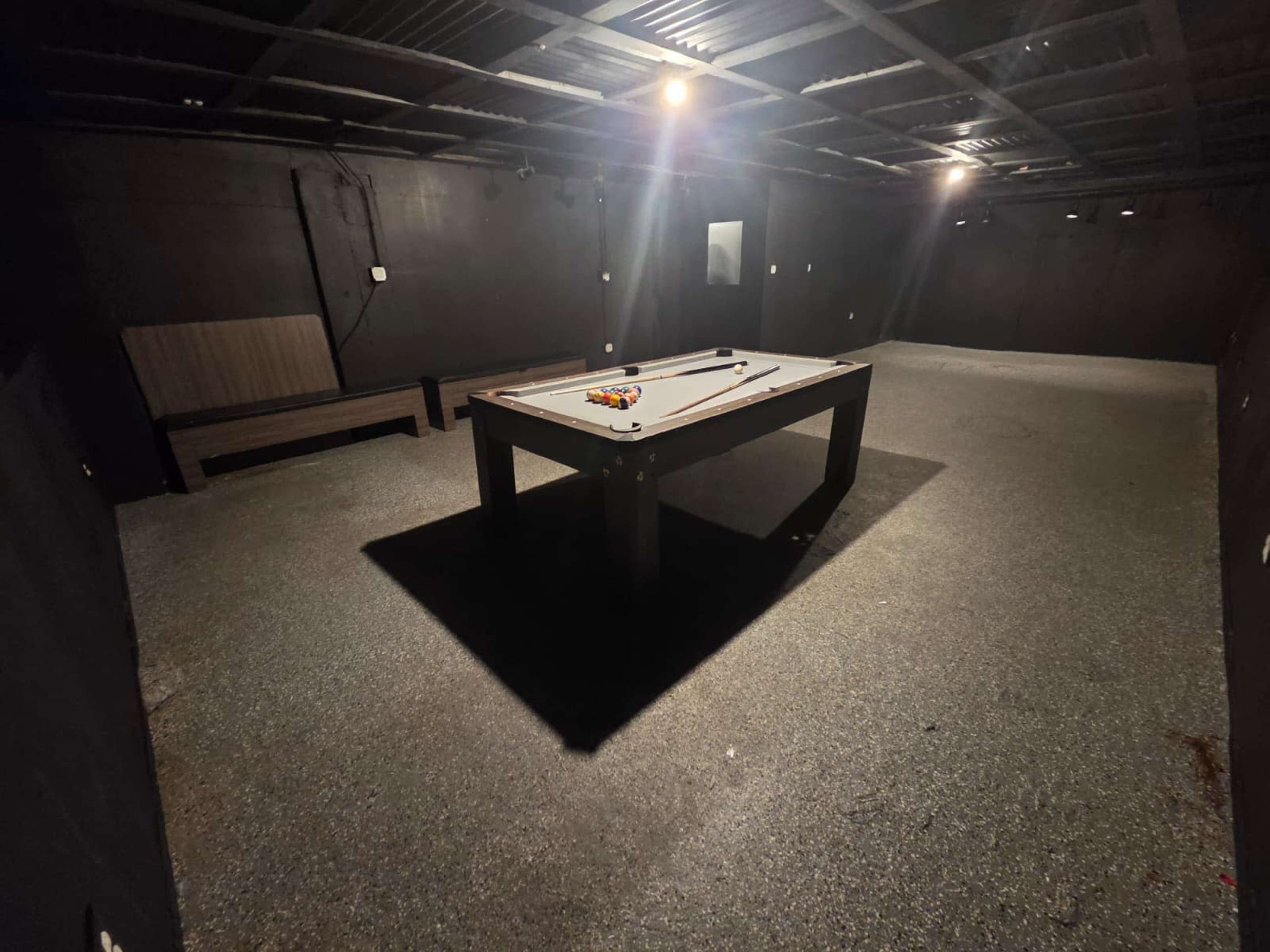 The image shows an empty billiard room with a pool table in the center and a bench against the wall.
