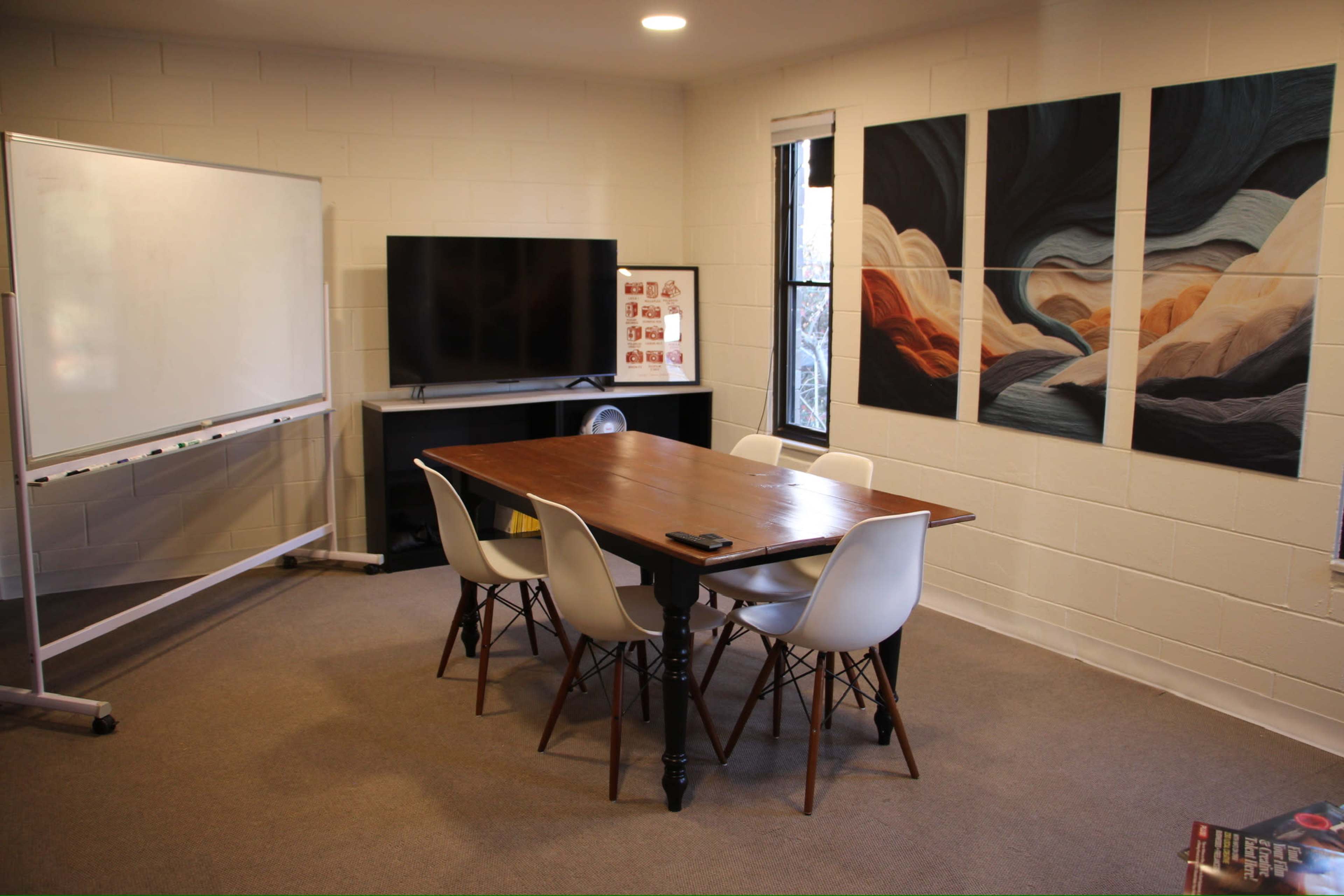 A meeting room is set up with a wooden table surrounded by white chairs, a television, a whiteboard, and abstract wall art.