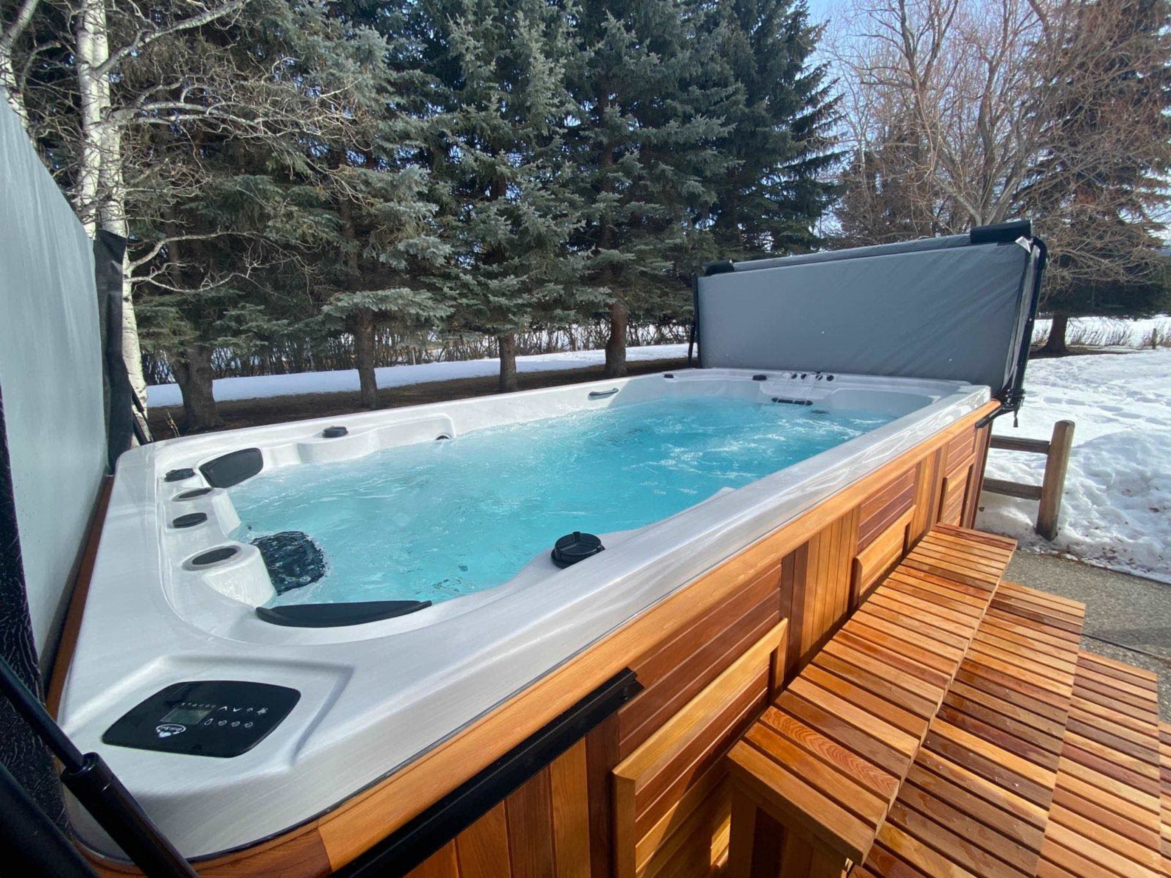 A rectangular hot tub with a wooden exterior and a covered top is situated outdoors near snow-covered ground and tall evergreen trees.