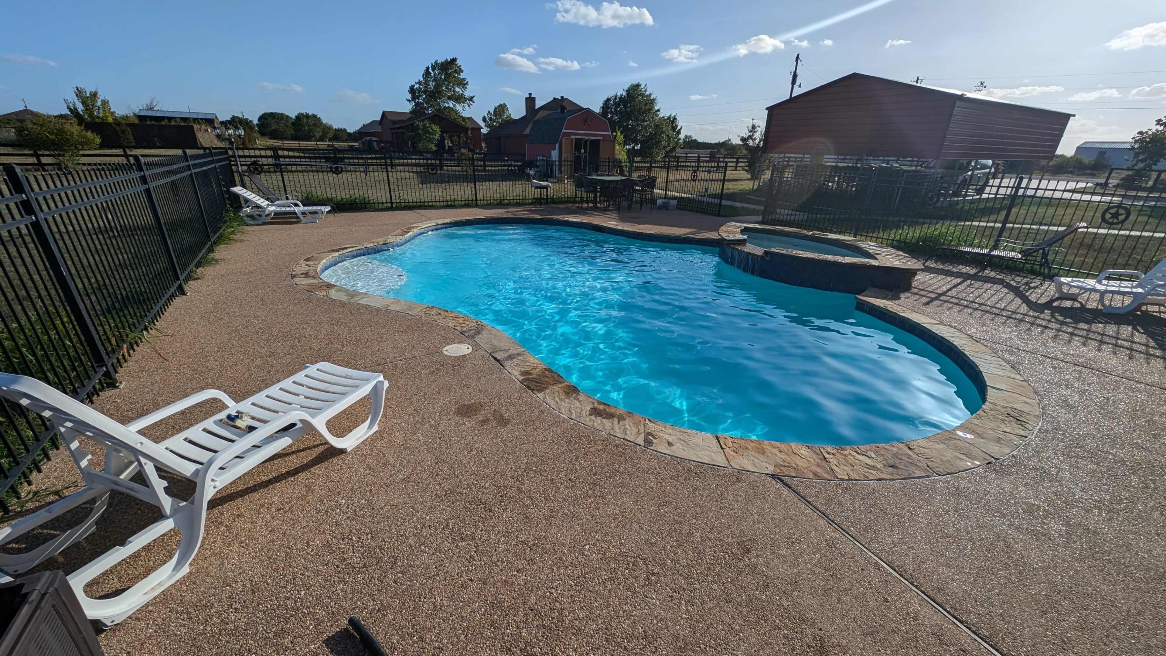 A swimming pool surrounded by a concrete deck and lounge chairs, set within a fenced backyard.