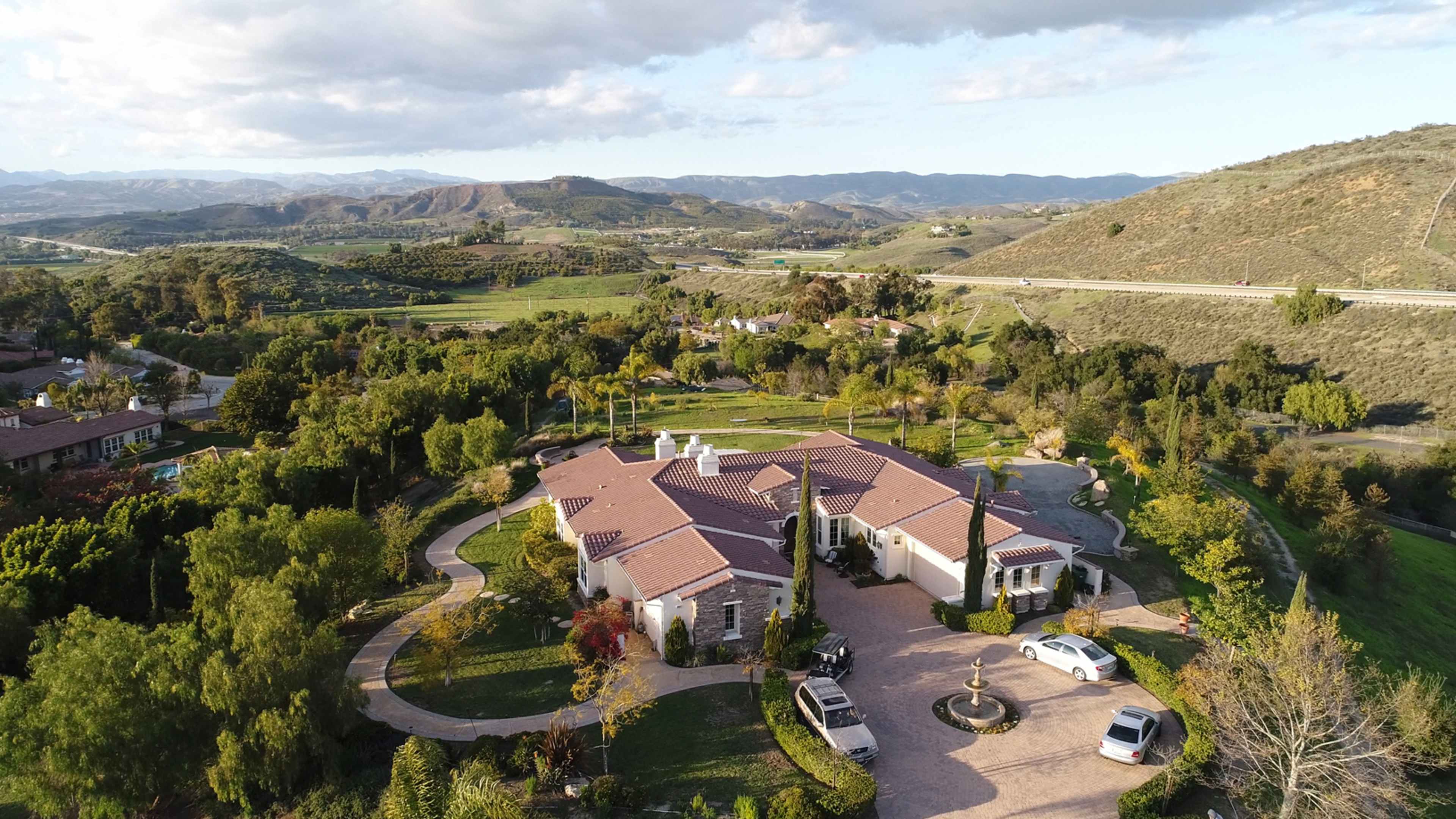 A large, sprawling house surrounded by green hills and trees, with several cars parked in the driveway.