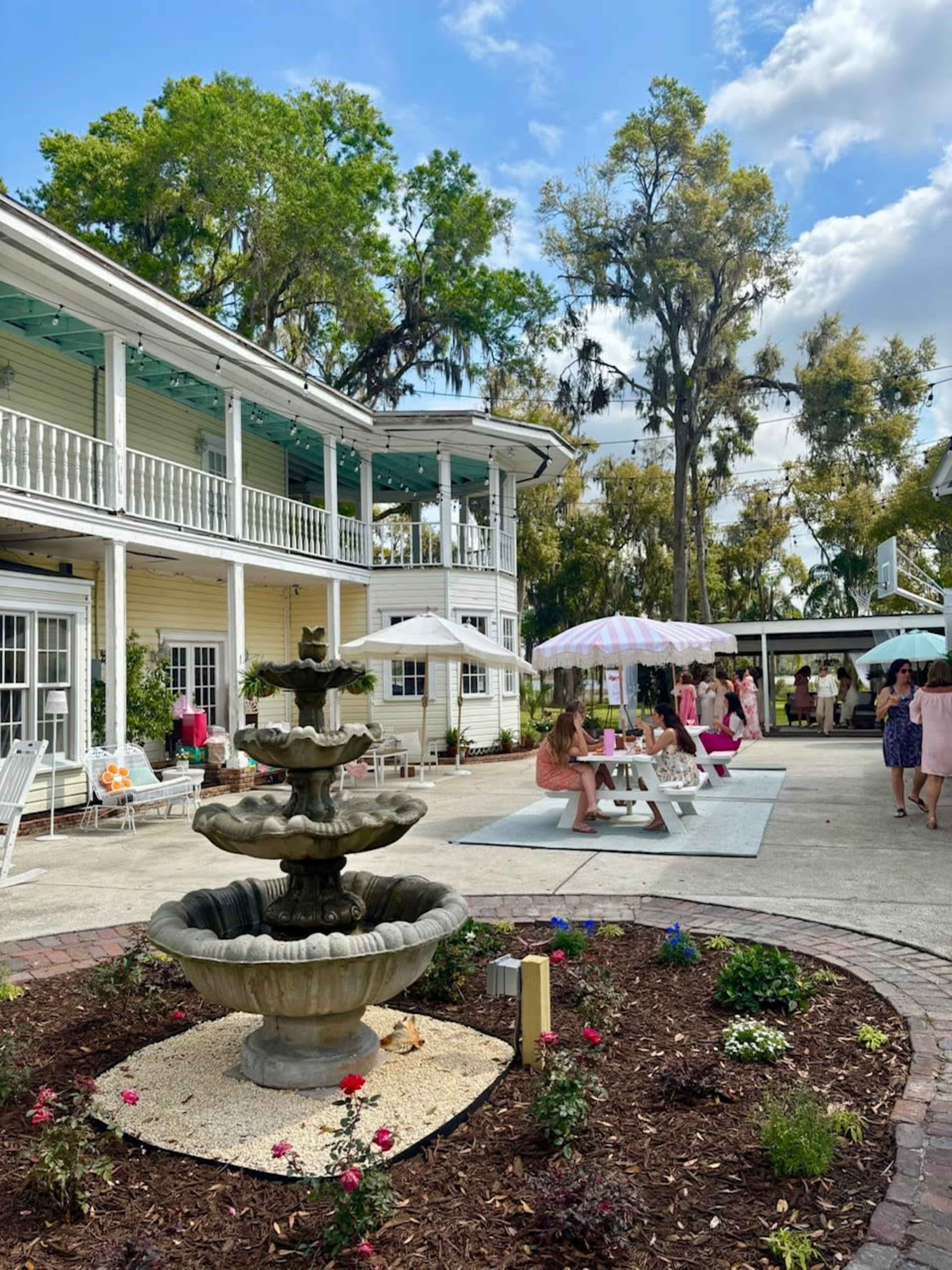 A courtyard features a multi-tiered fountain surrounded by flowering plants, with a historic building and people socializing under umbrellas in the background.
