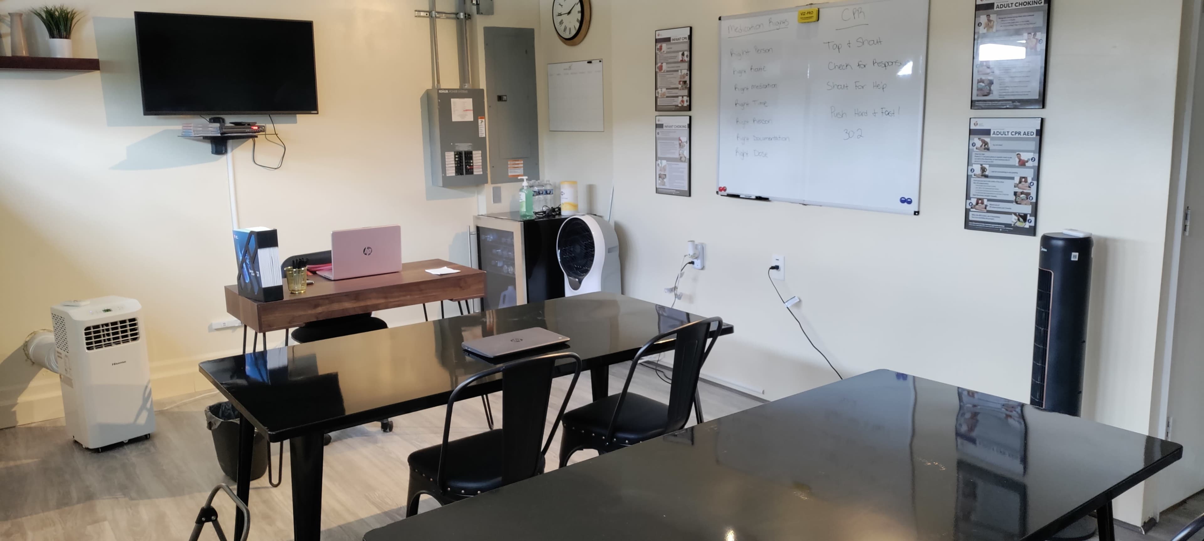 A small classroom or meeting space featuring a wall-mounted television, a whiteboard with notes, a desk with a laptop, and two black tables with chairs.