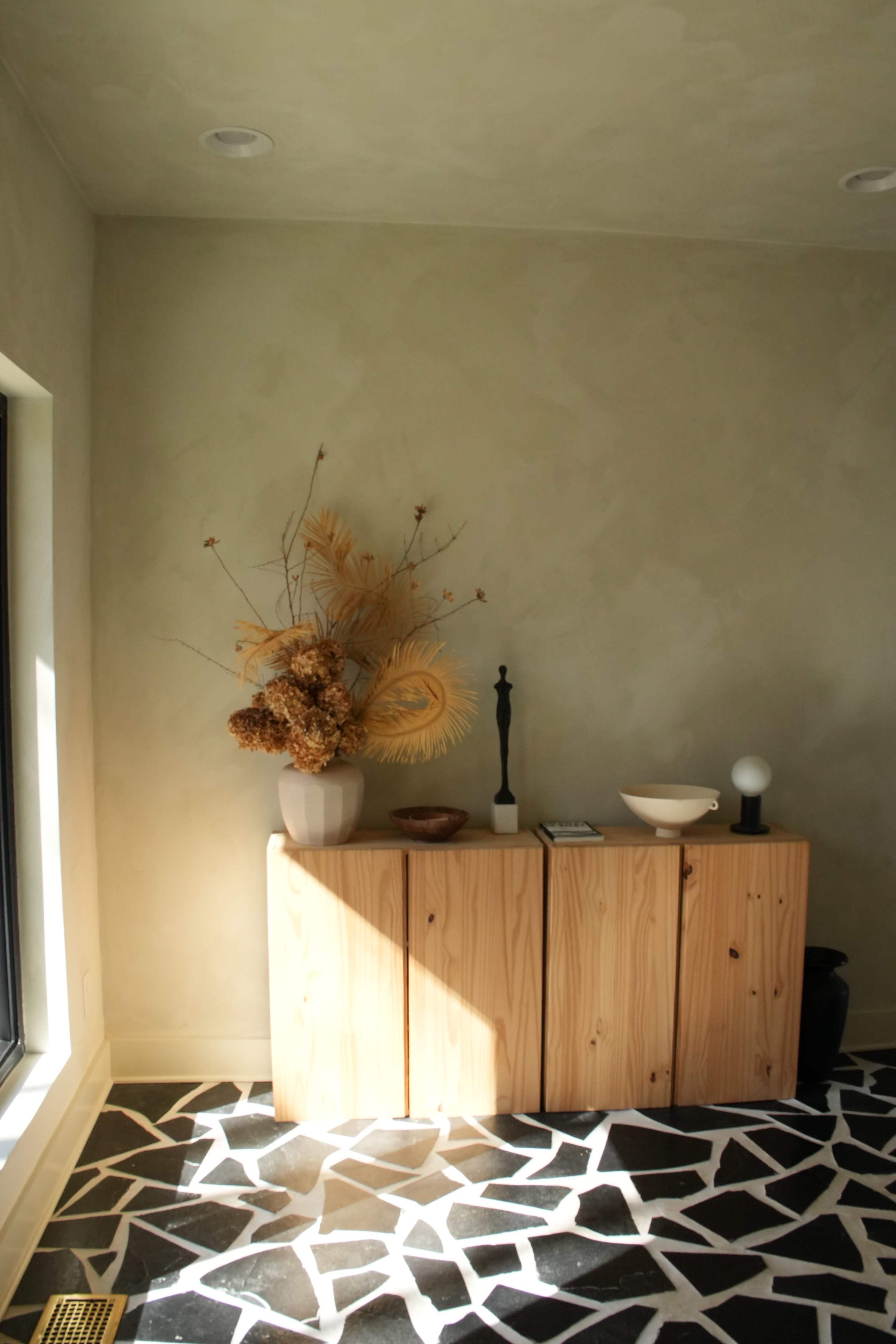 The image features a minimalist interior with a light-colored wall, a wooden cabinet topped with a decorative vase, and a striking geometric black and white floor.