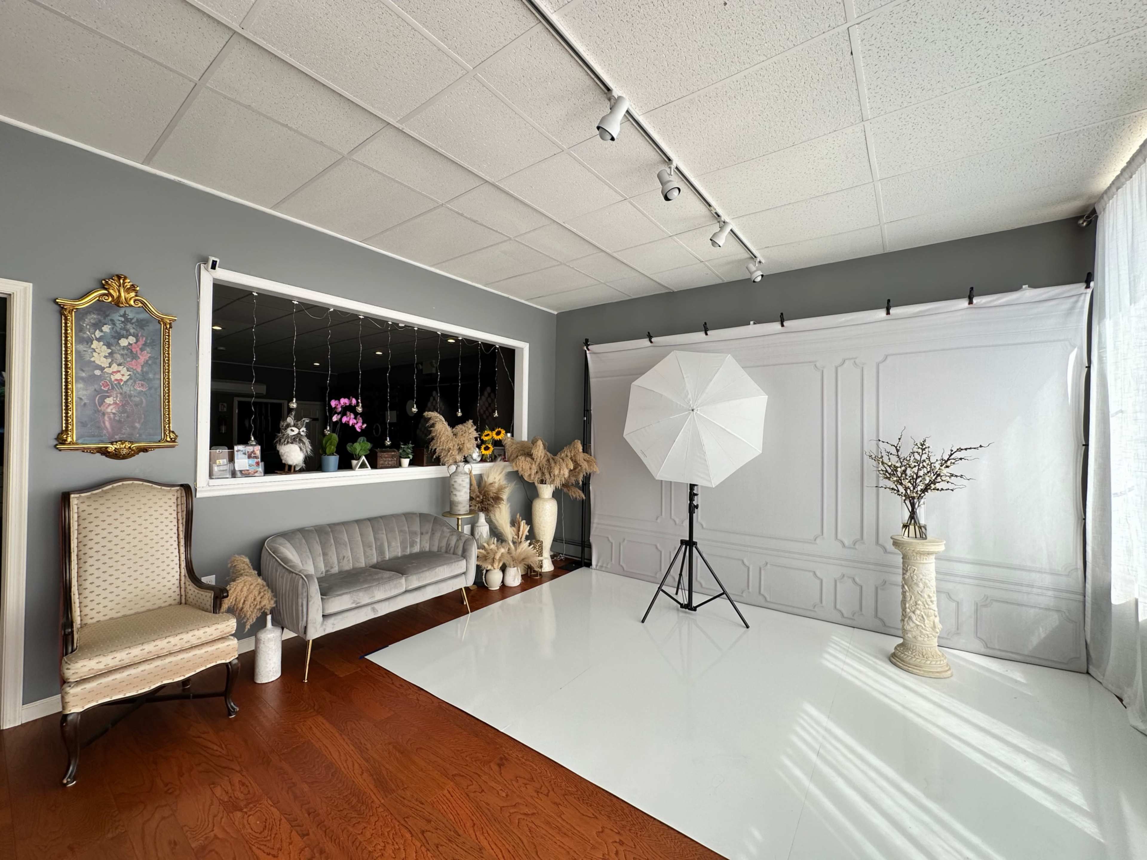 A photography studio featuring a gray wall, white backdrop, soft lighting equipment, and elegant furnishings including a vintage chair and decorative vases.