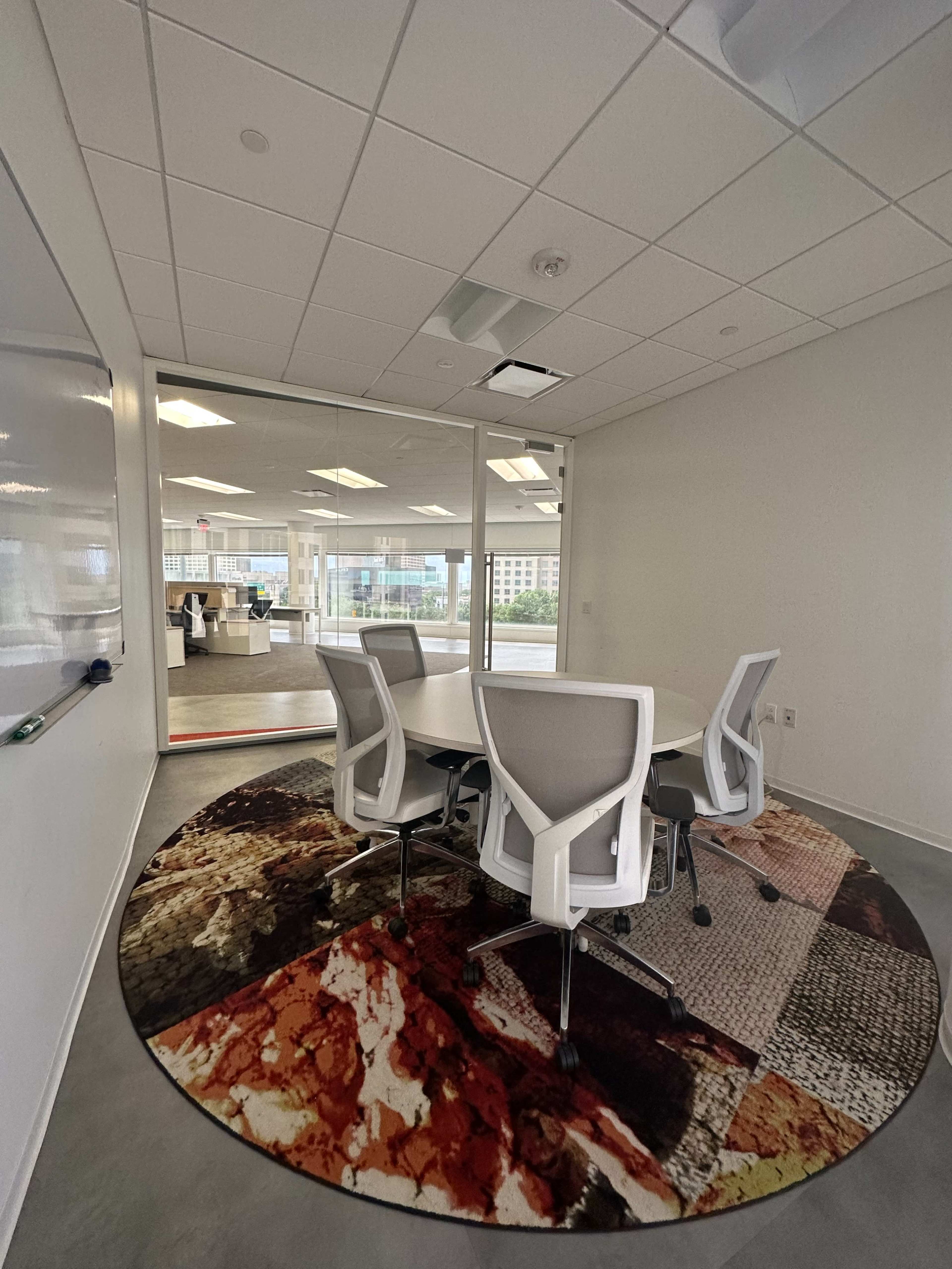 A modern conference room features a round rug under a large table surrounded by white chairs, with glass walls offering a view of an office space beyond.
