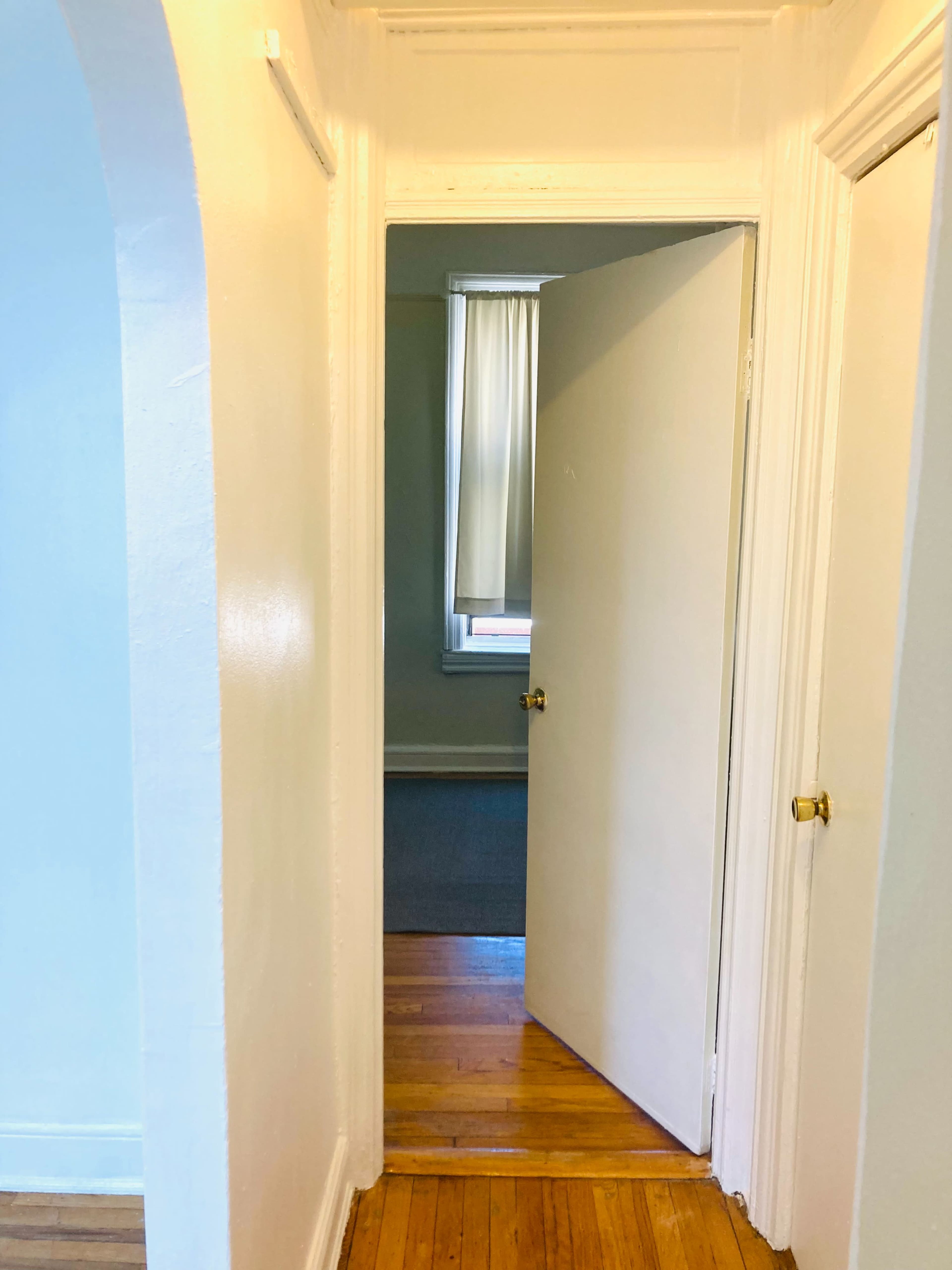 A hallway with a white wall leads to an open door, revealing a room with a window covered by a light curtain.