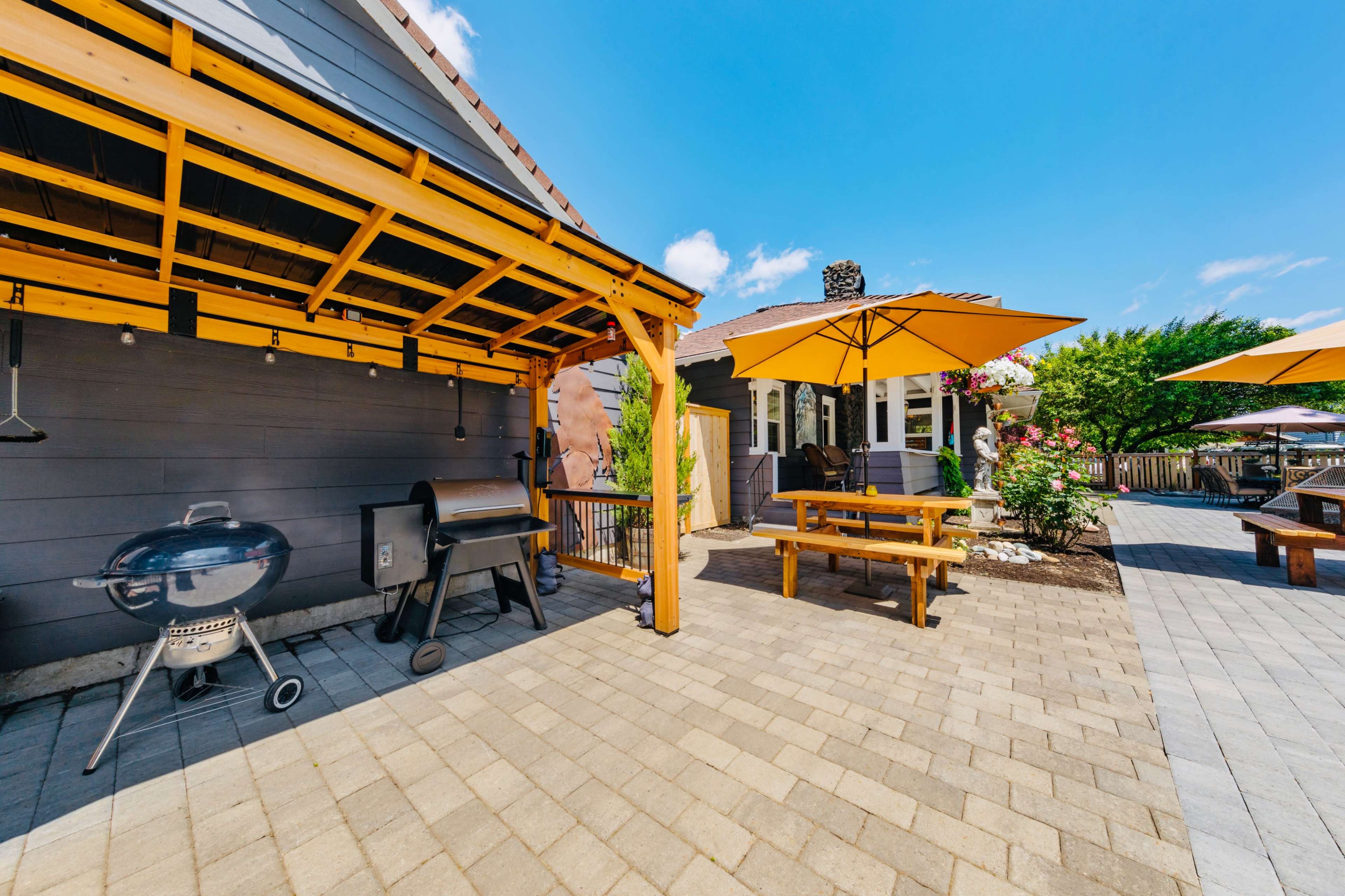 The image shows a patio area with a grill, a picnic-style table, and yellow umbrellas, surrounded by a well-maintained yard and a house in the background.