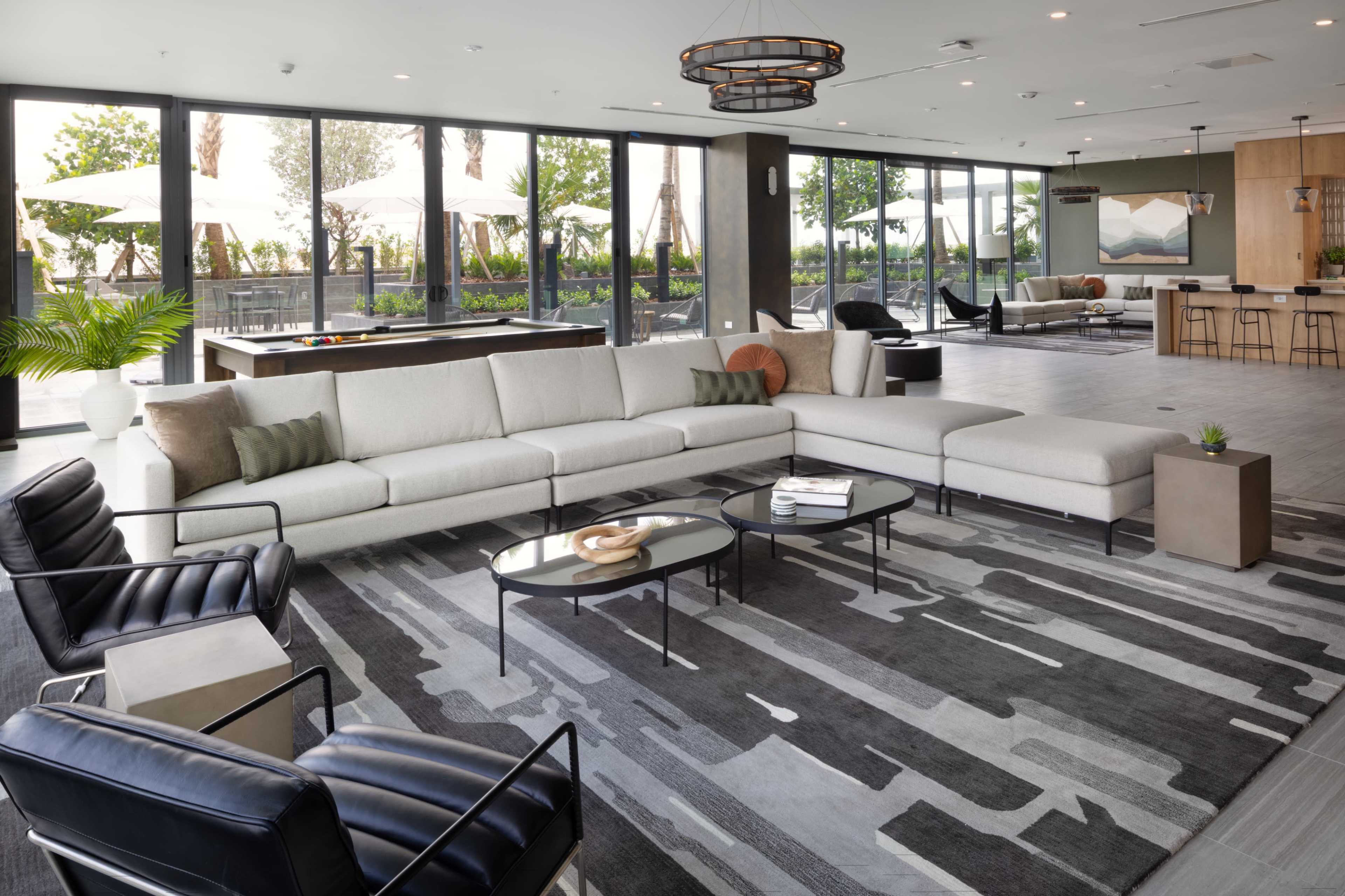 A modern lounge area features a large sectional sofa, two black chairs, a coffee table, and a pool table, with large windows offering views of an outdoor space.