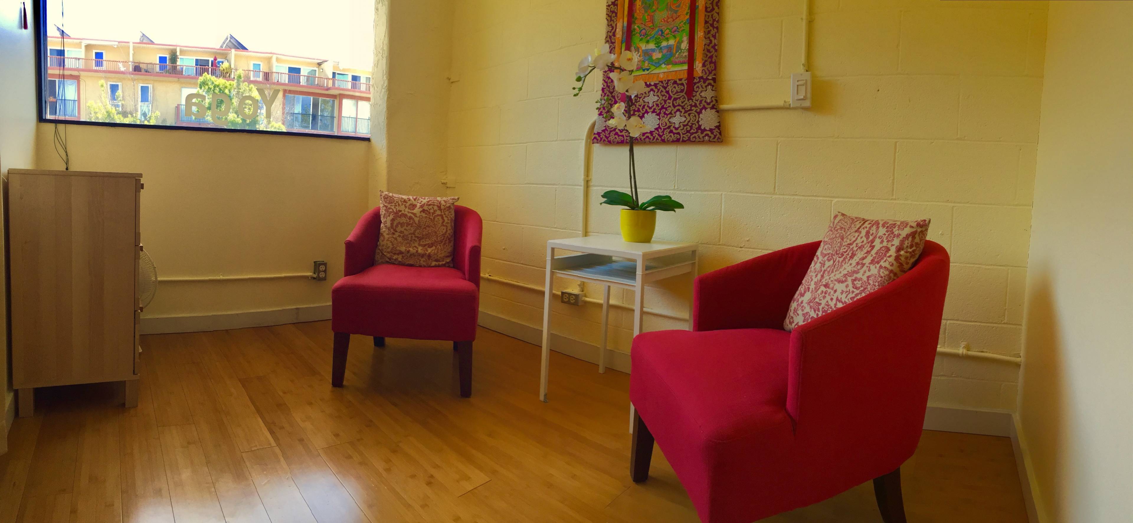 A bright room with two pink chairs, a small white table with a potted plant, and a window revealing a building outside.