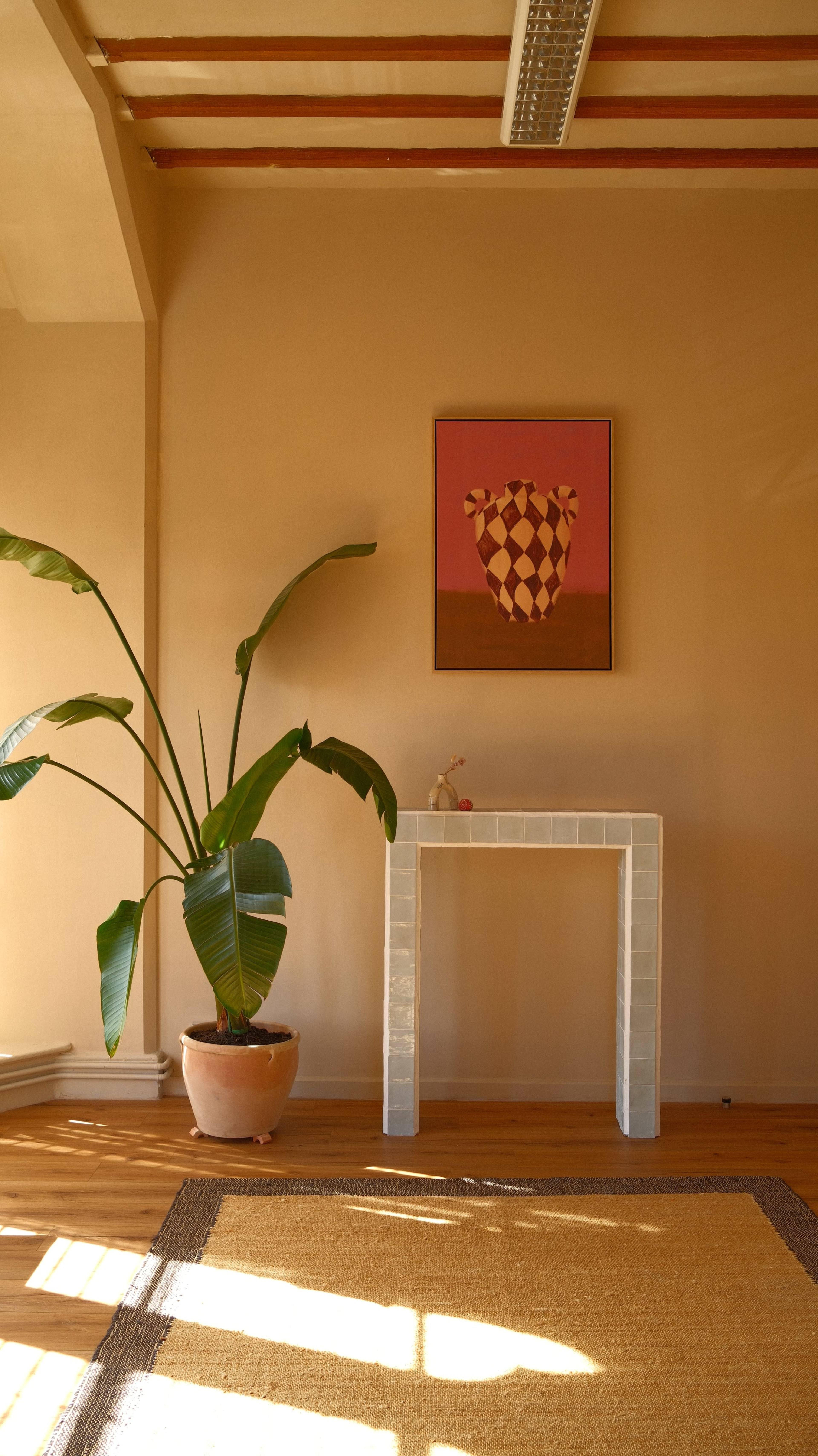 A potted plant stands next to a square, white table that holds a small decorative item, under a framed artwork on a beige wall.