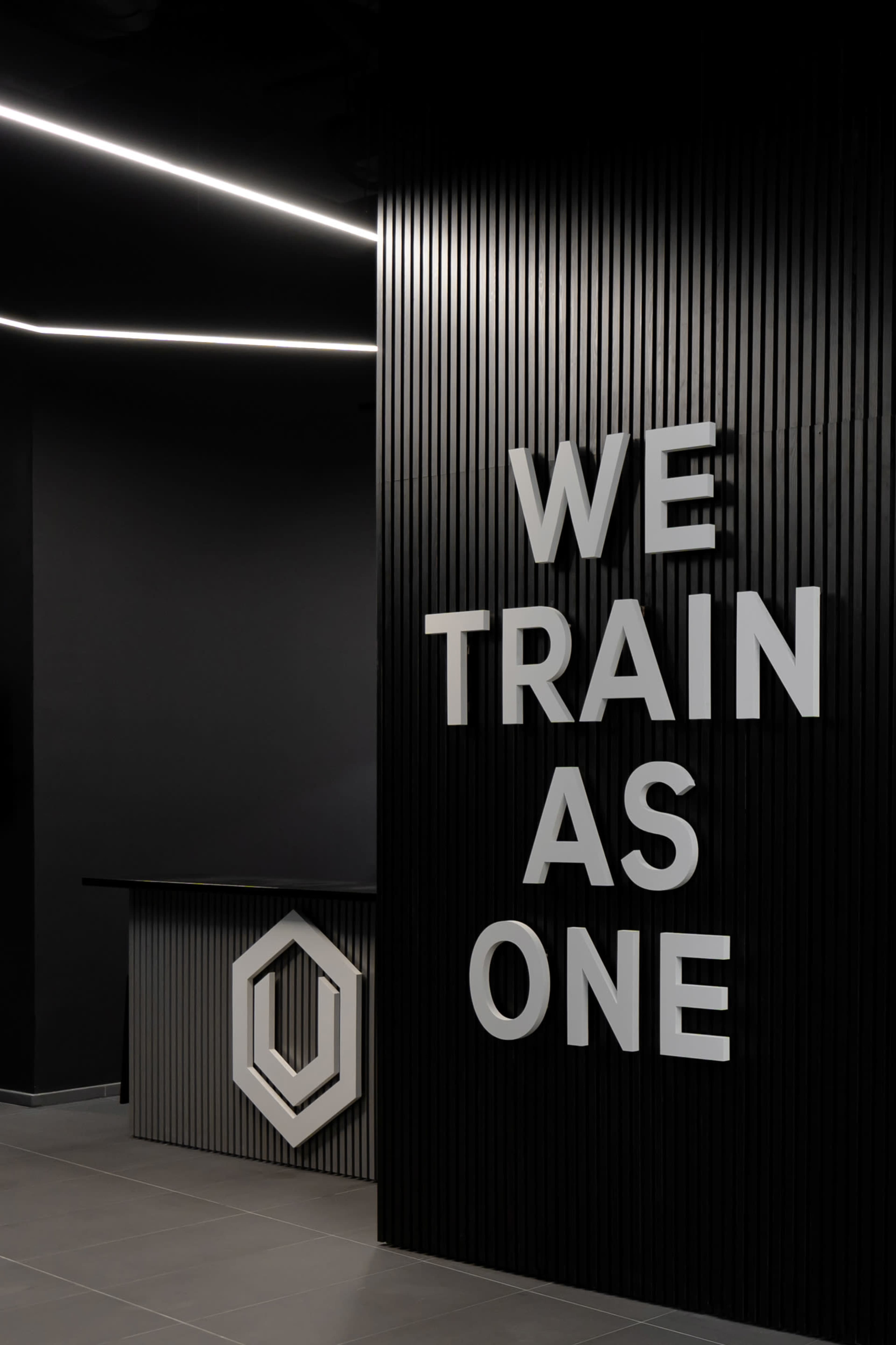 A modern reception area with a black wall featuring the phrase "WE TRAIN AS ONE" in large, white letters beside a geometric logo.