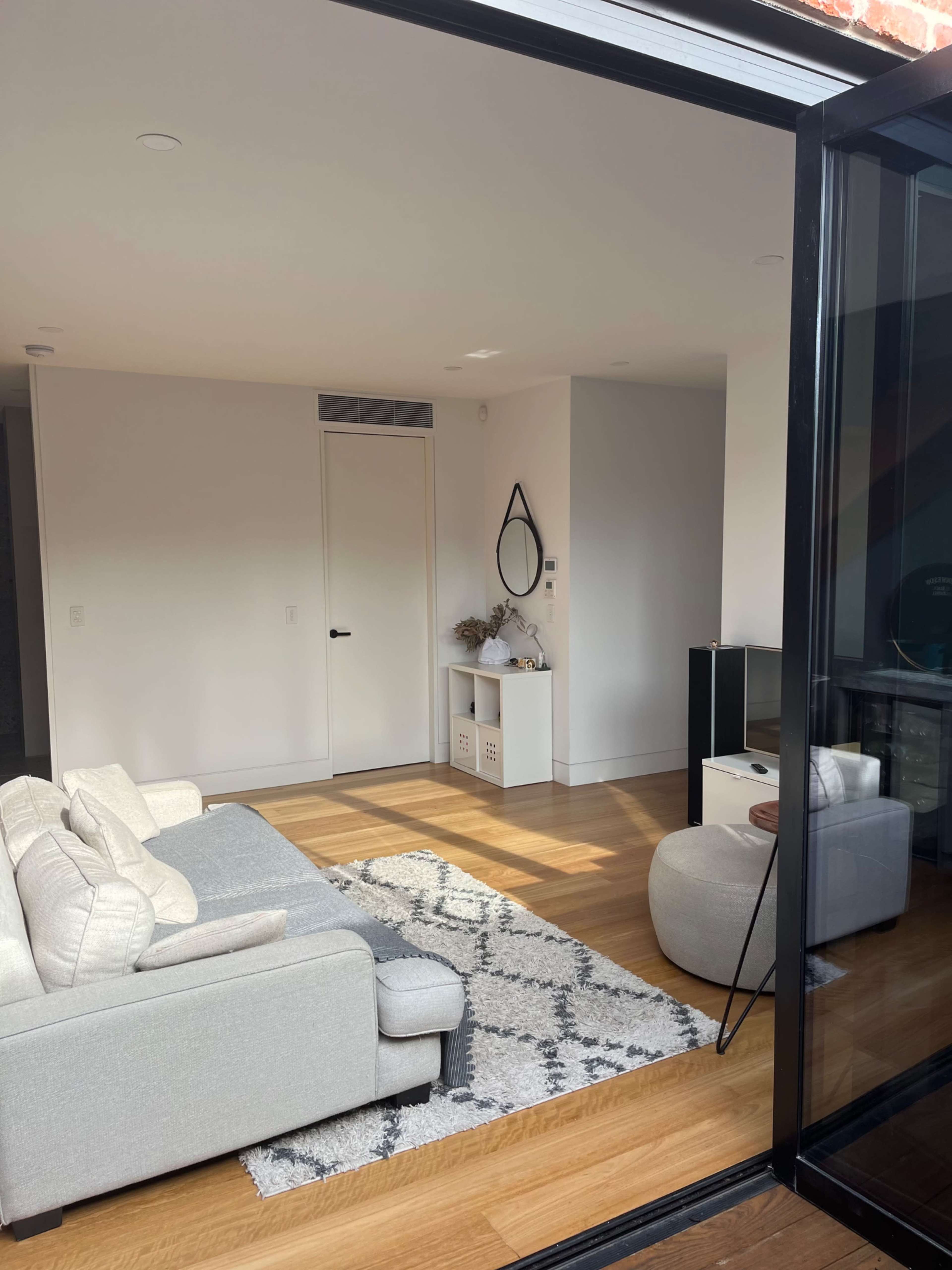 A light-filled living room features a gray sofa, a geometric rug, and a TV unit against a wall with a door and a mirror.