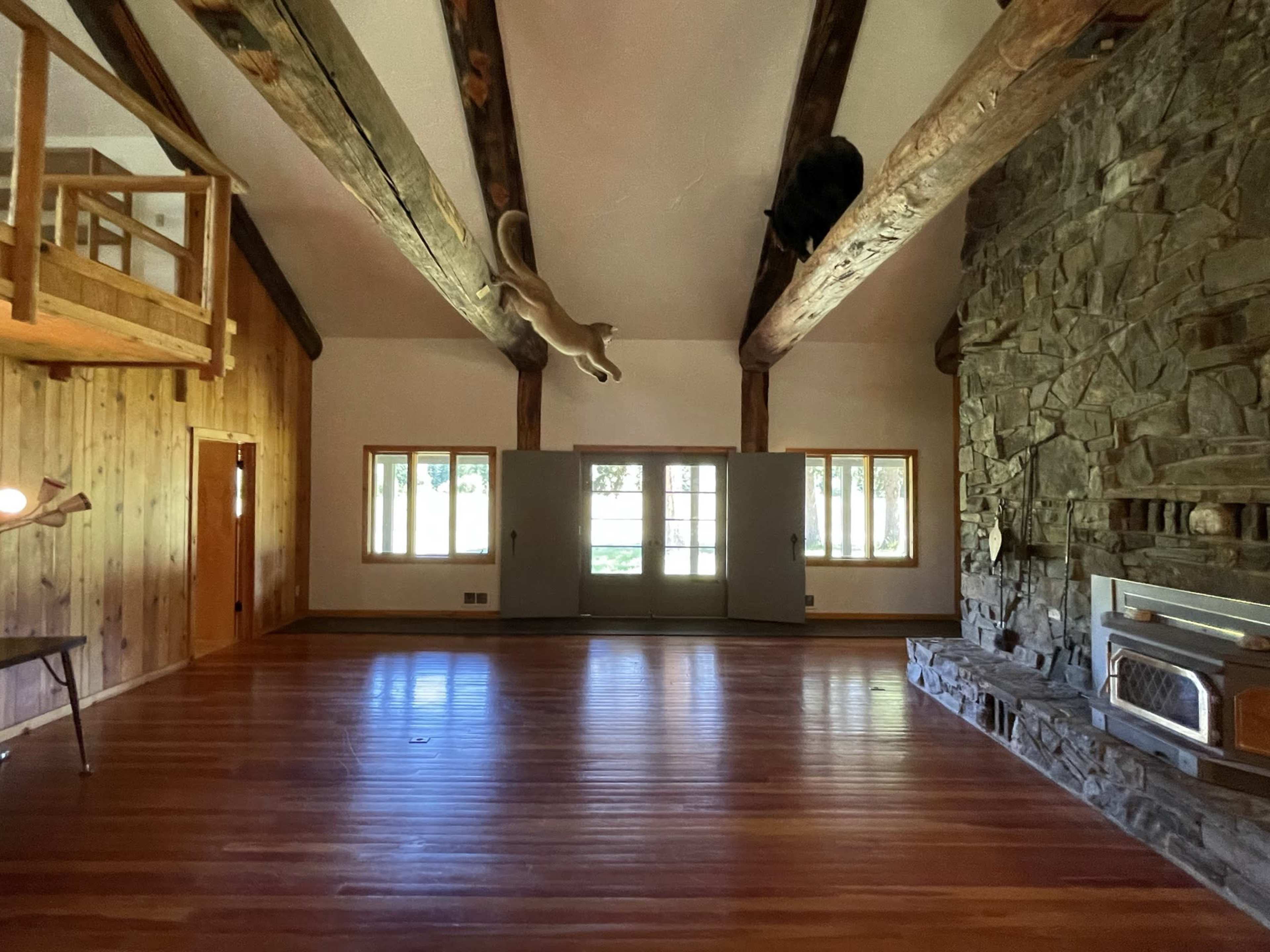 A spacious room with high, wooden beam ceilings, a stone fireplace, and large windows overlooking a garden.
