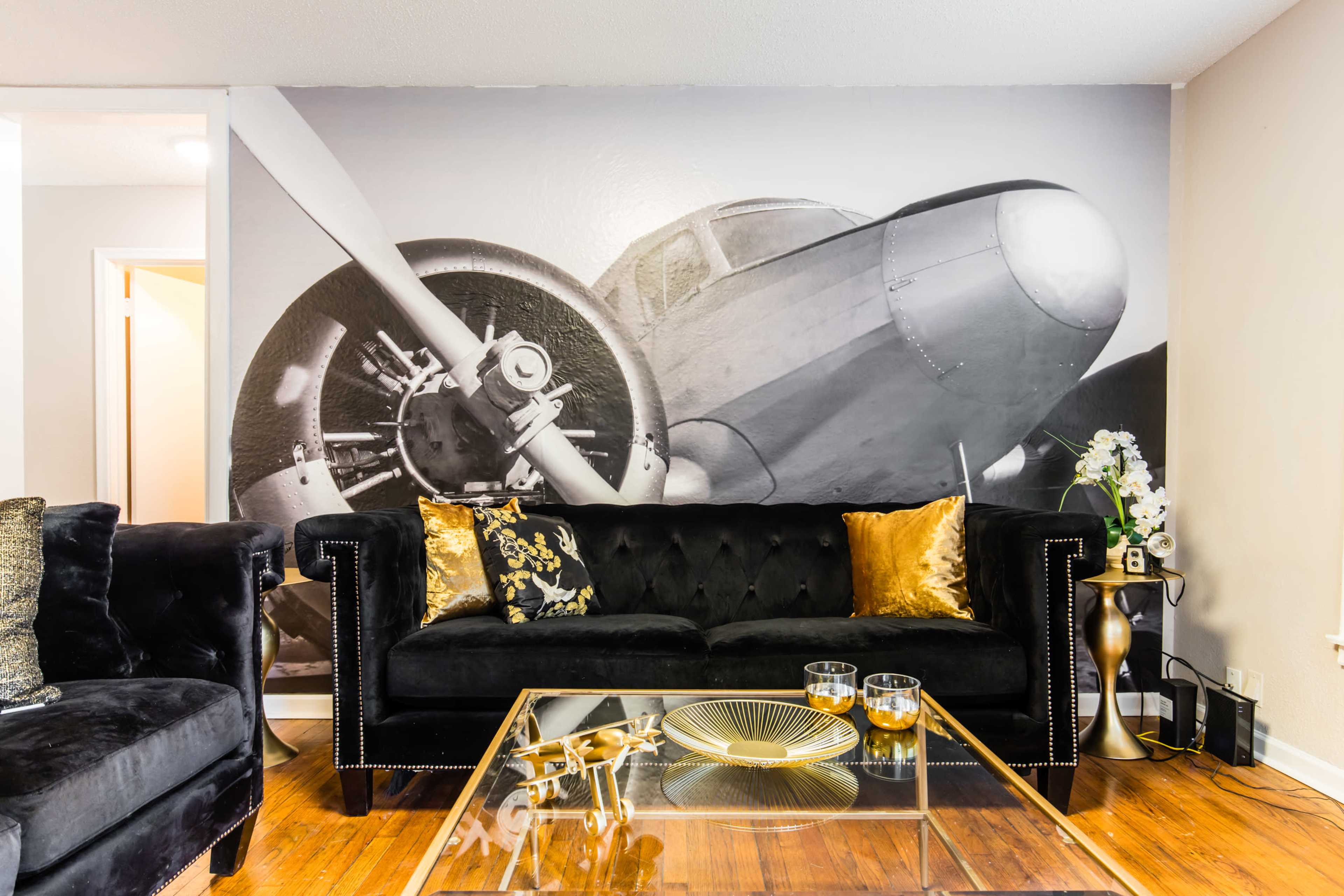 The living room features a black velvet sofa adorned with gold pillows, facing a glass coffee table in front of a large black-and-white wall mural of an airplane.