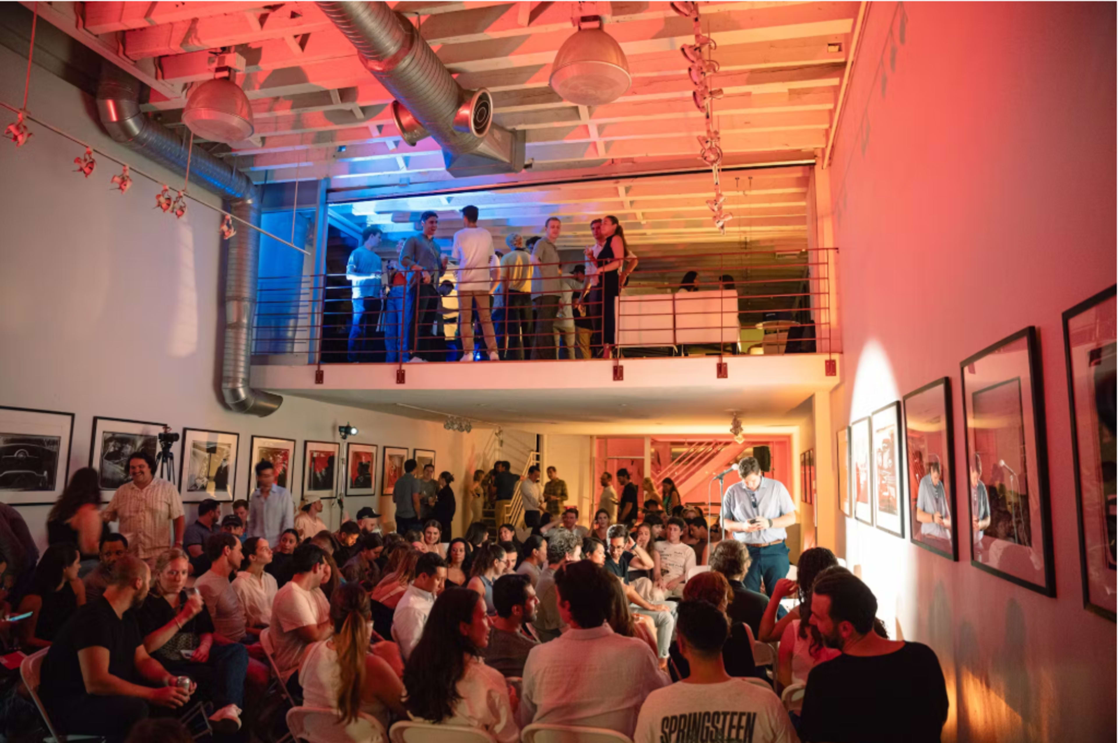 A large group of people is gathered in a well-lit room with artwork on the walls, while an individual speaks on a stage in front of an audience seated on chairs.