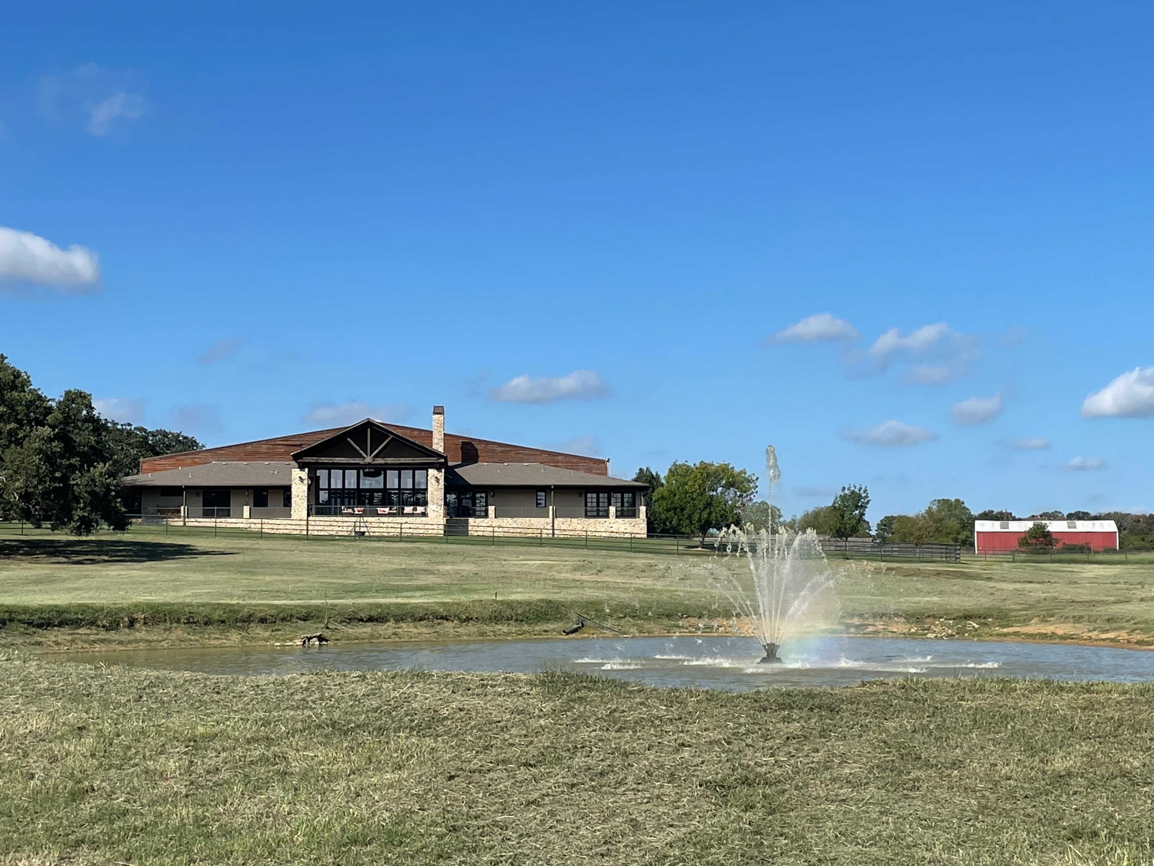 A large, modern building with a stone facade overlooks a pond featuring a fountain in a grassy area under a clear blue sky.