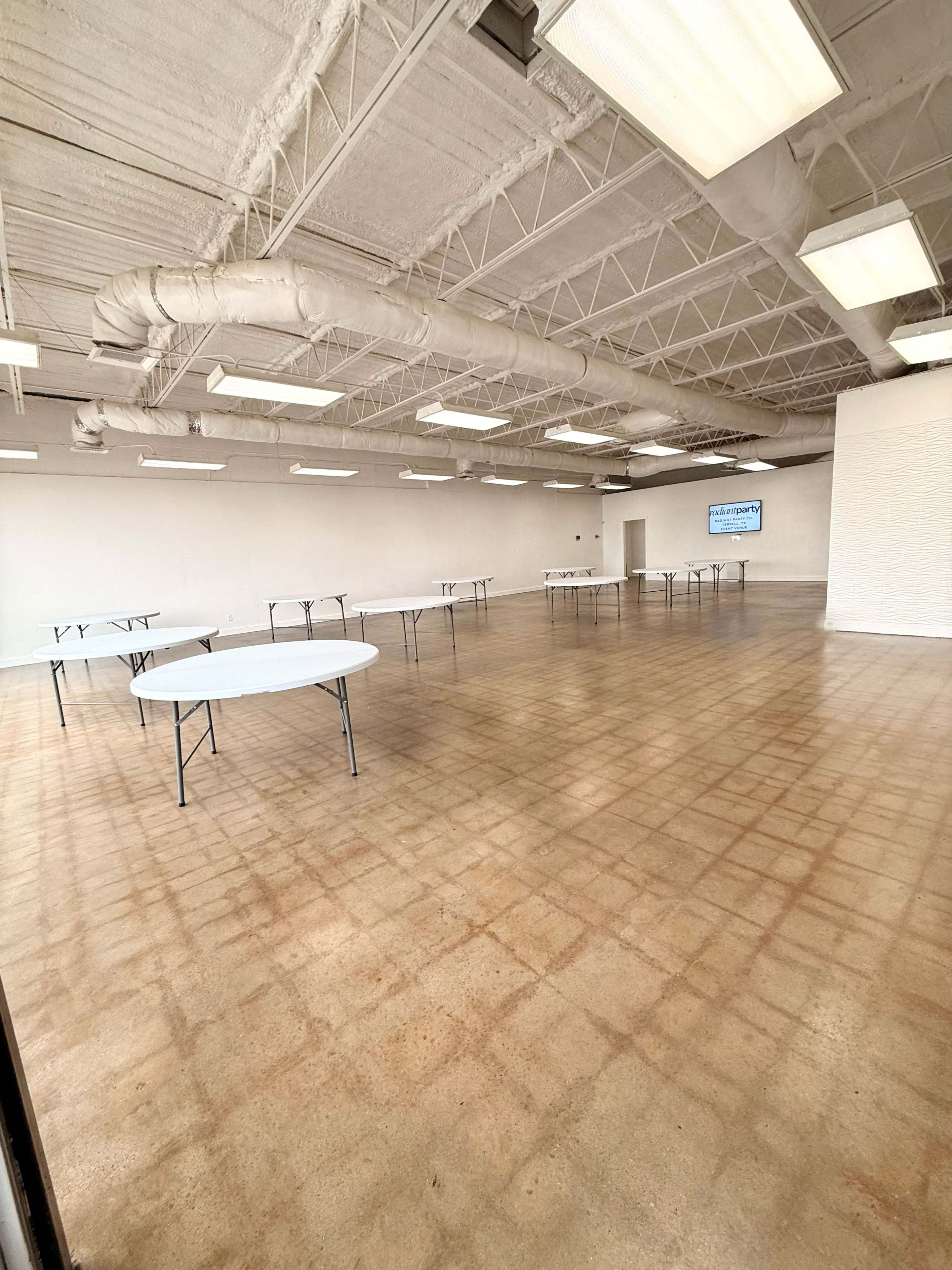 An empty room featuring multiple round folding tables arranged throughout the space, with a white wall and exposed ceiling.